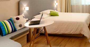 Bedroom with a laptop on a small table; bed with pillows, small sofa, light wood floor.
