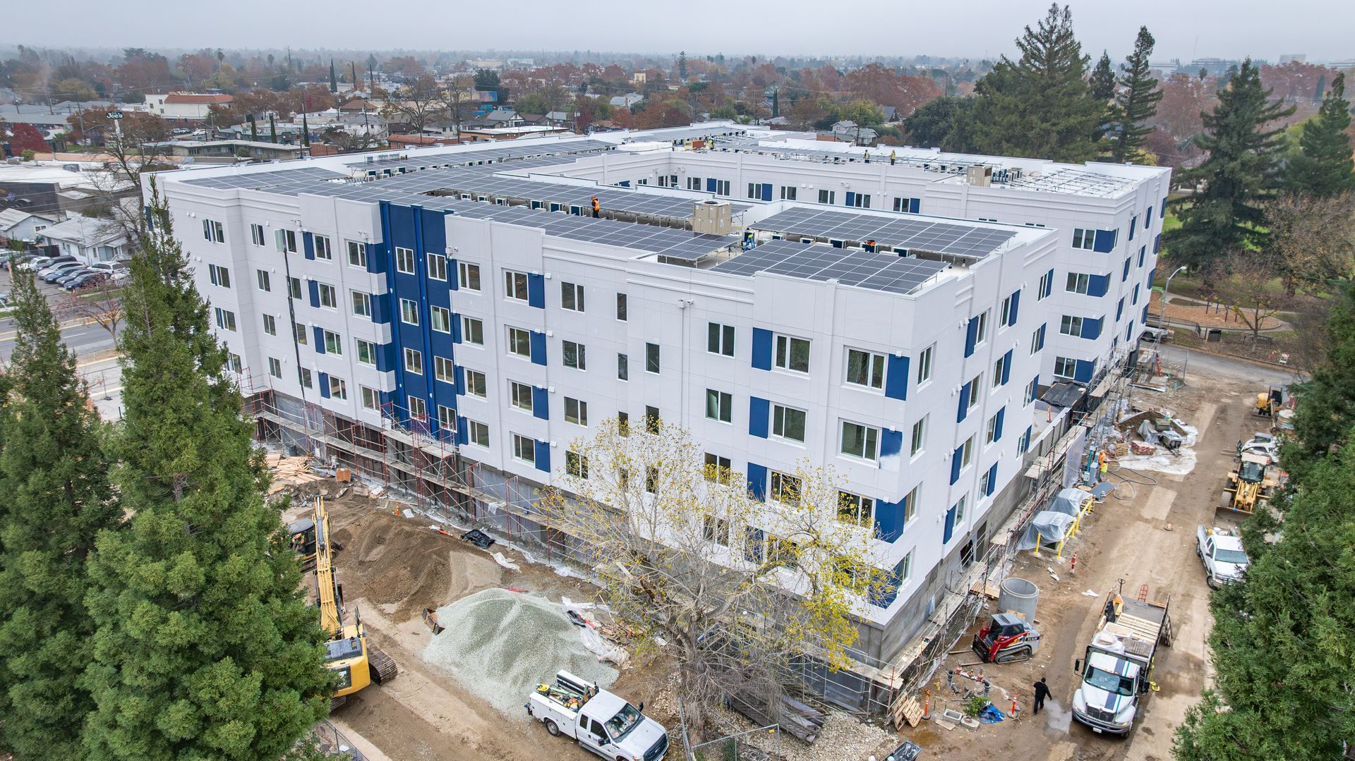 Aerial view of a white apartment building with blue accents, surrounded by trees and construction equipment