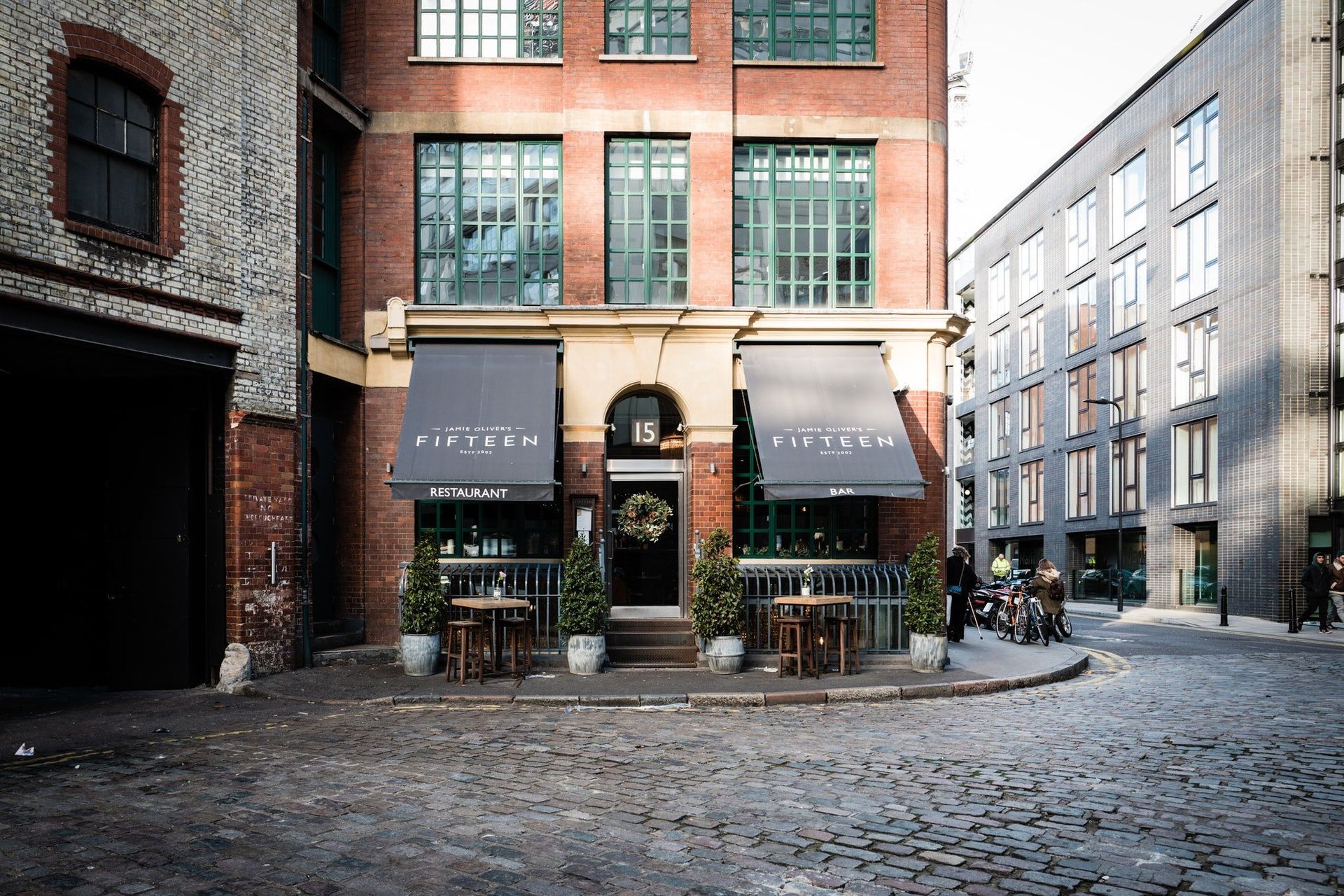 Brick storefront on a cobblestone street with awnings and outdoor tables, beside a narrow lane.