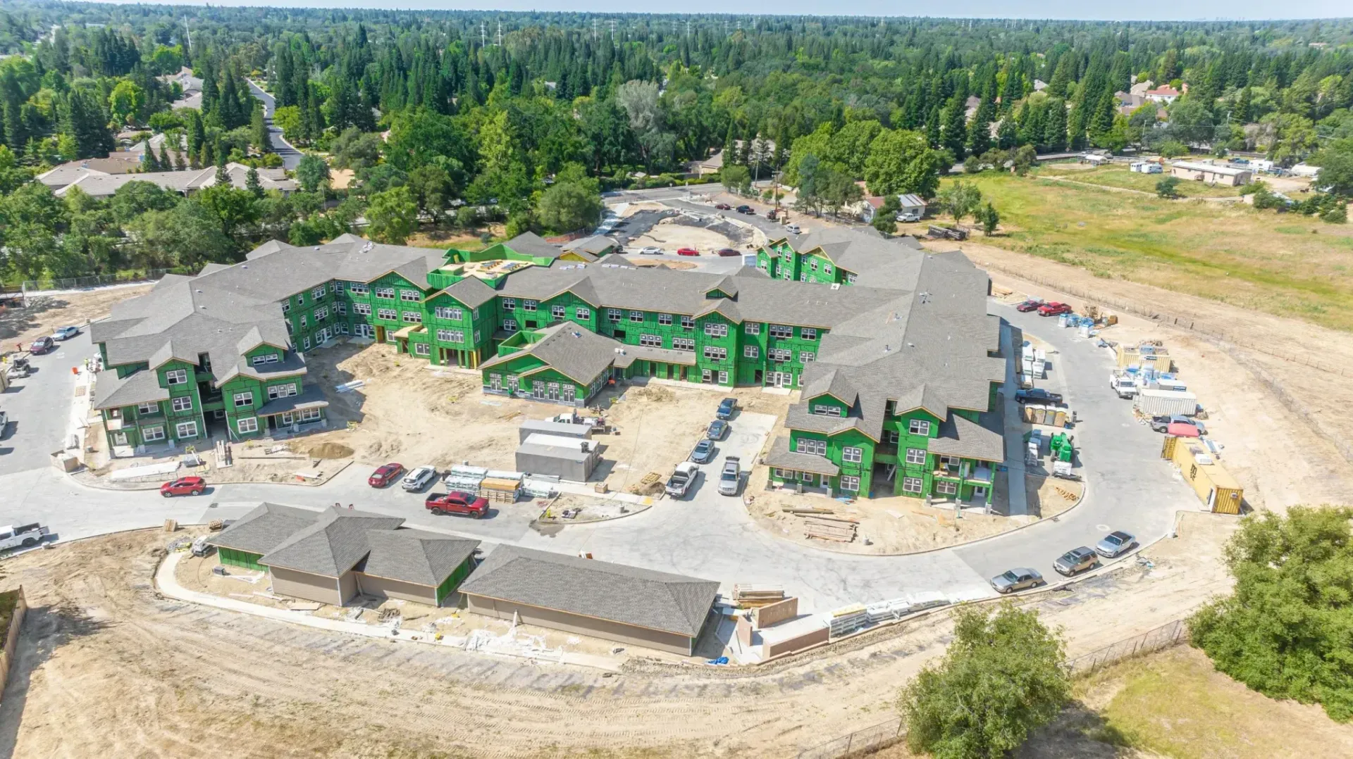 Aerial view of a large construction site with gray roofs, green tarps, and surrounding trees.