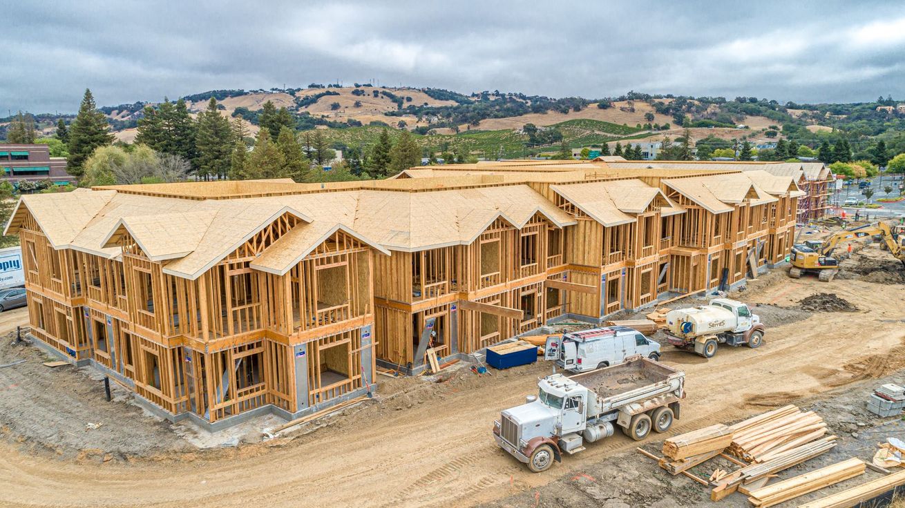 Large wooden apartment buildings under construction with trucks and lumber on a dirt site, hills in background