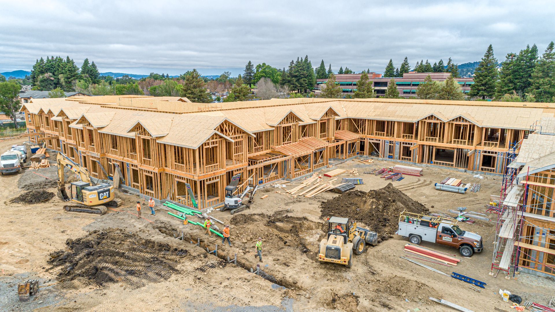Large wooden building under construction at a muddy site with trucks and scaffolding beneath cloudy skies