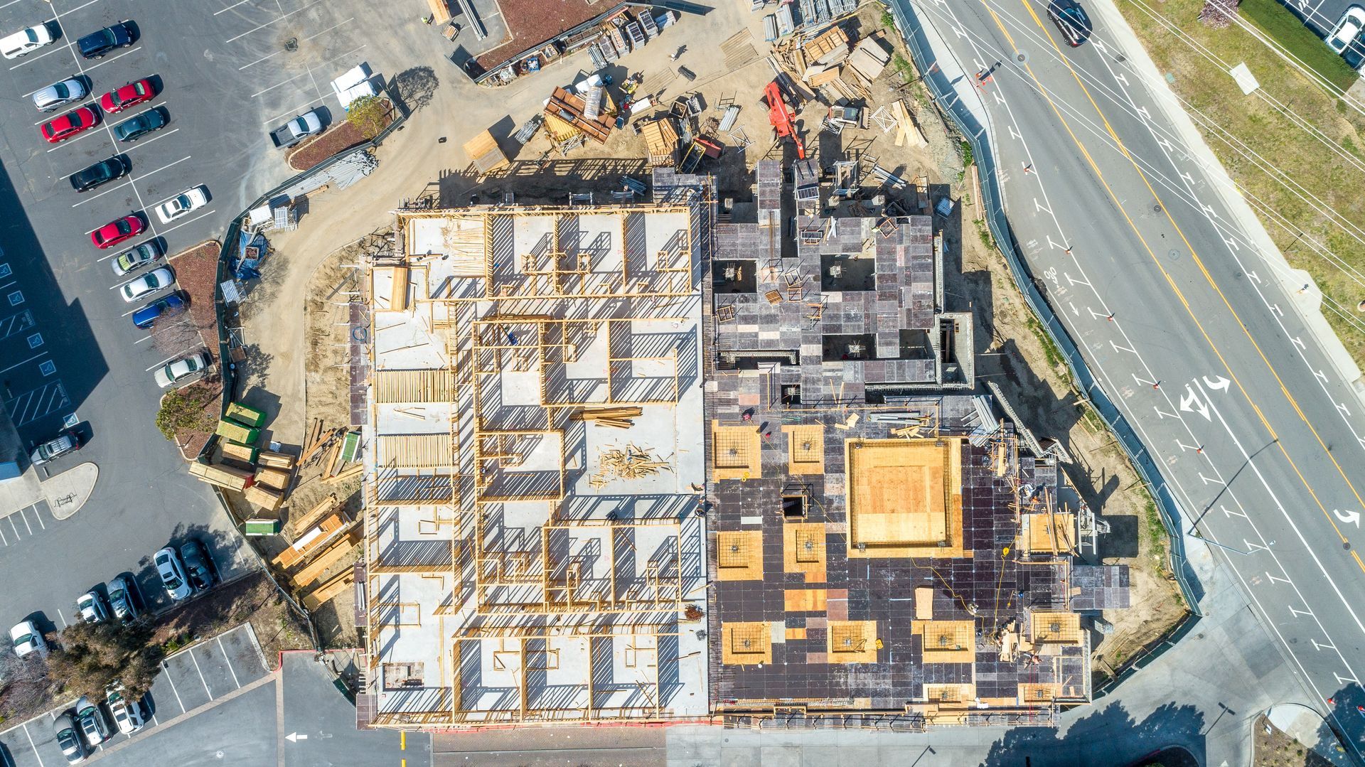 Aerial view of a partially built building site with exposed framing, construction materials, and nearby parking lots.