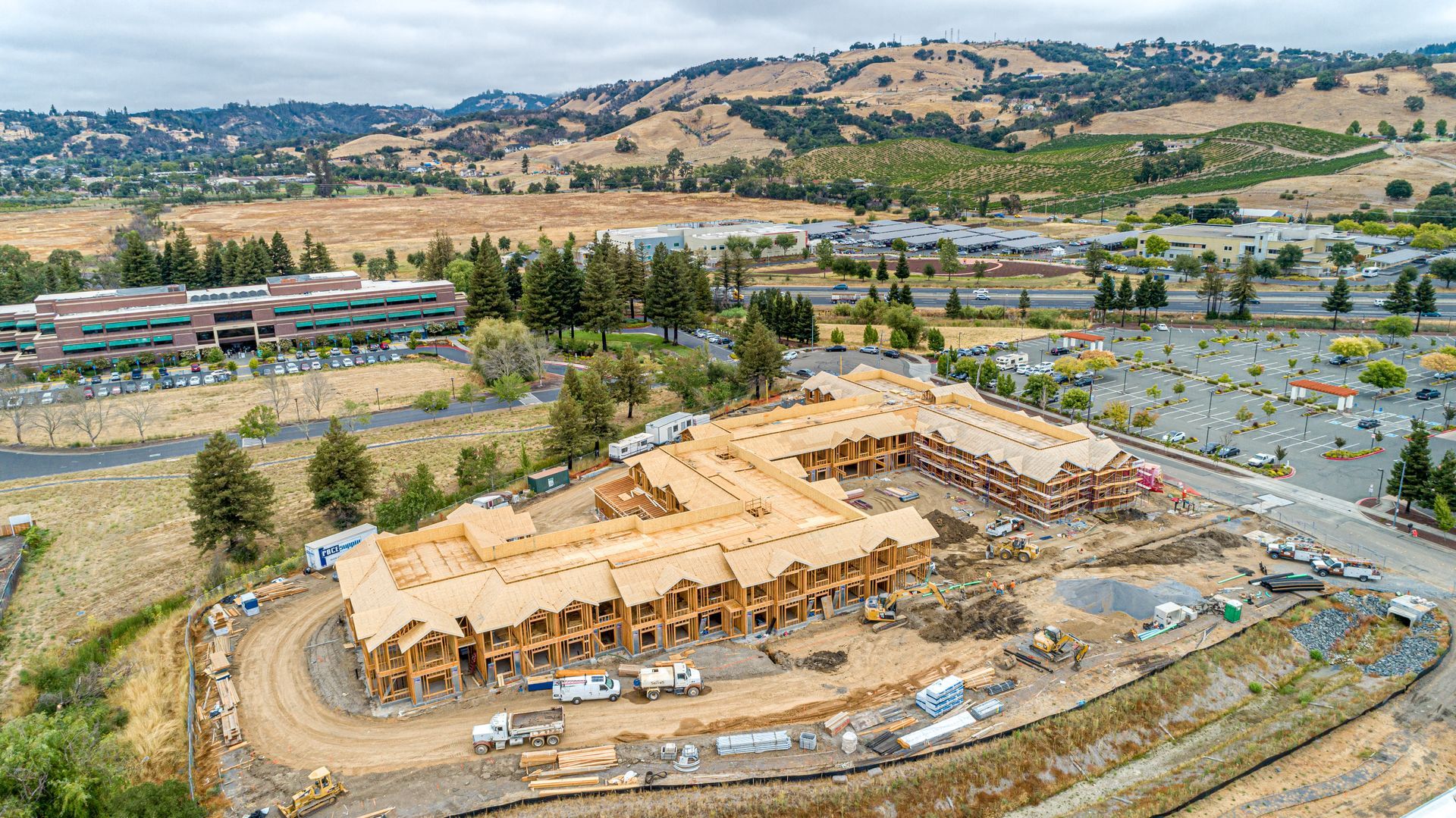 Aerial view of a large tan building under construction beside parking lots and hills under a clear sky