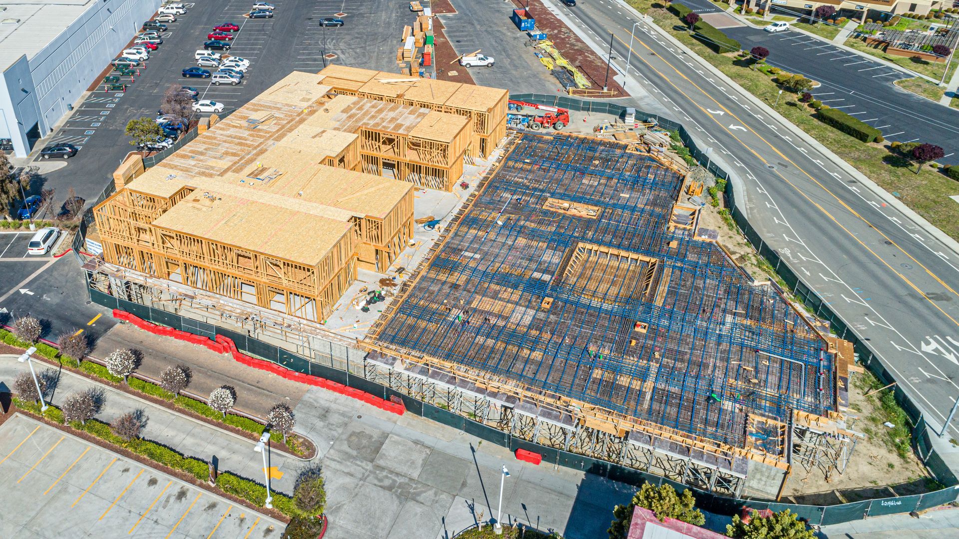 Aerial view of a large building under construction beside a road and parking lot, with exposed wood framing and dark roofing.