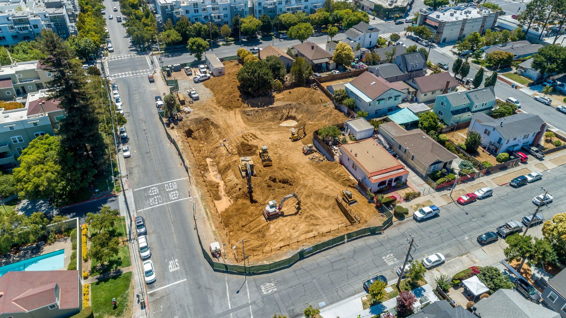 Aerial view of a construction site on a city block, bordered by roads and homes with parked cars nearby.