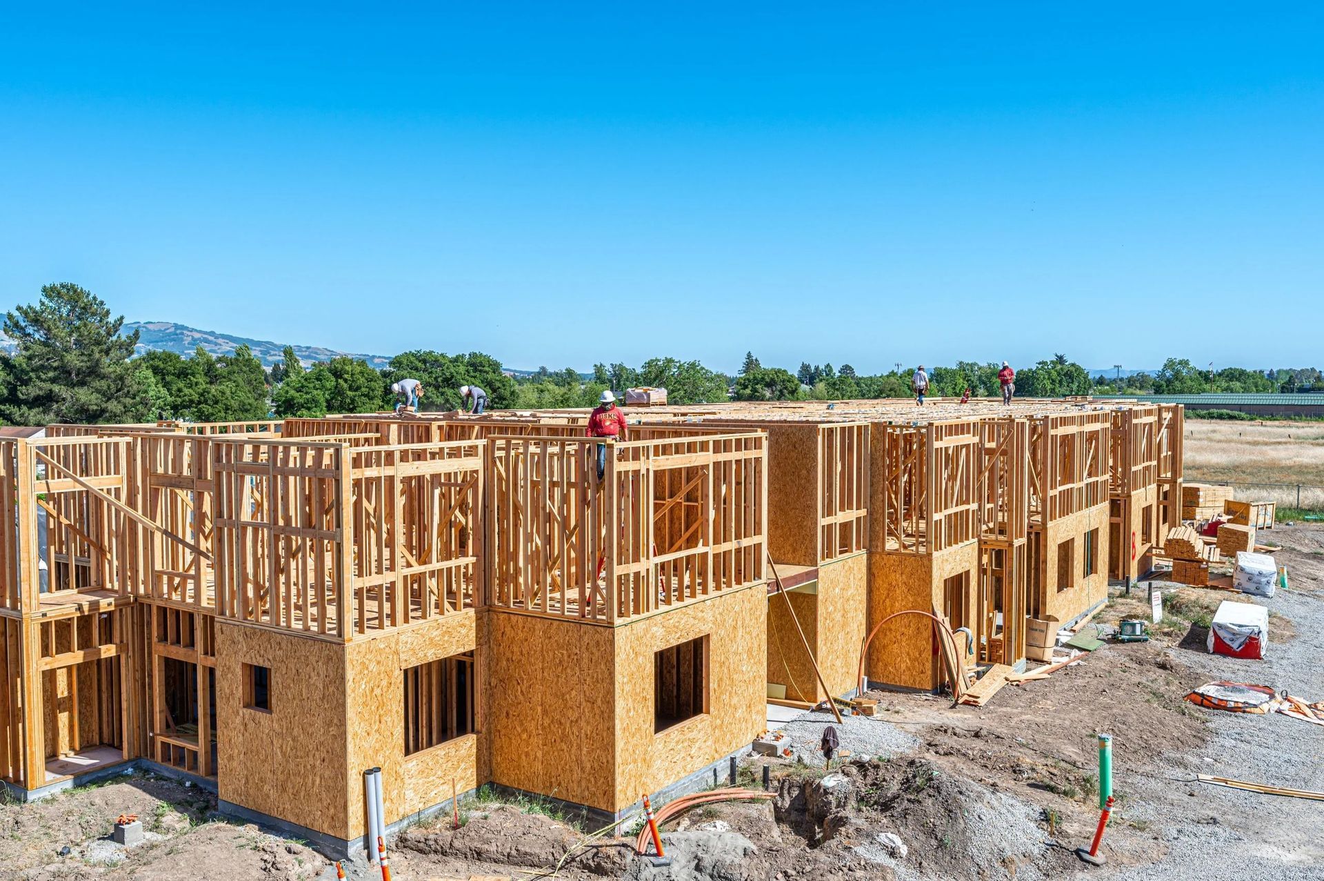 Construction workers framing a long, multi-unit residential building with wooden walls against a clear blue sky.