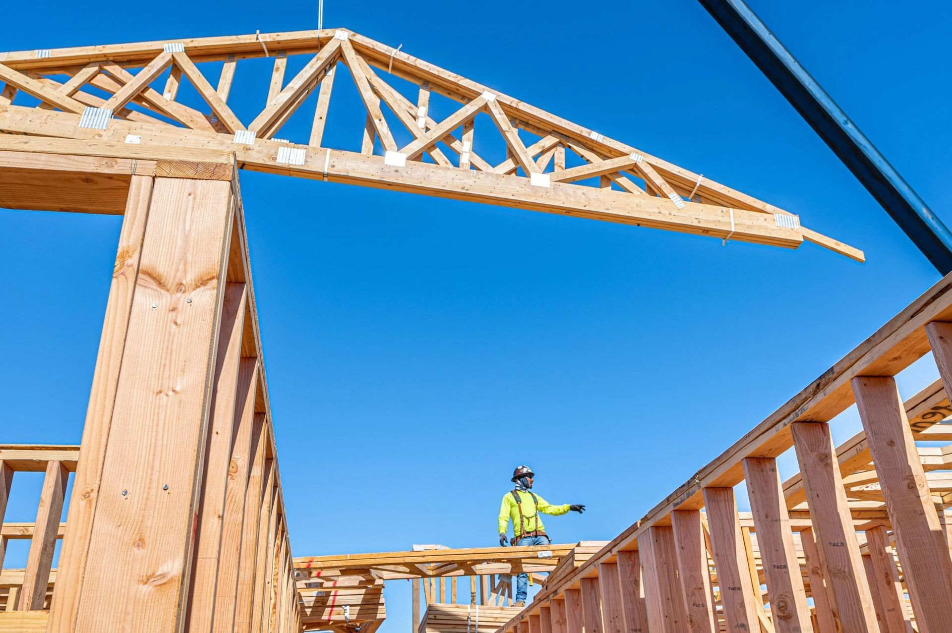 A construction worker in a high-visibility vest directs a crane lifting a wooden roof truss onto a building frame.