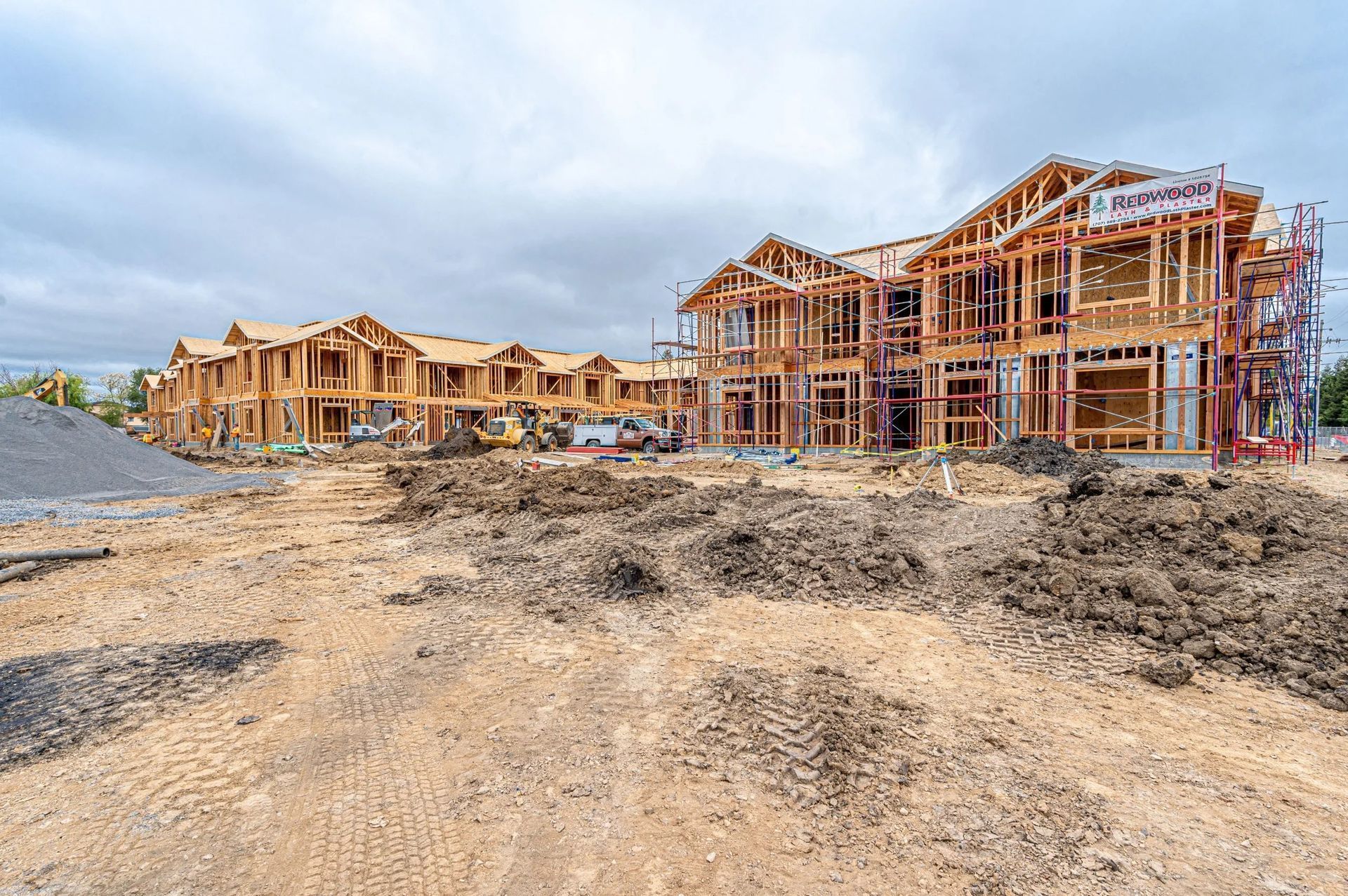 Two large wooden apartment buildings under construction sit on a dirt lot under a cloudy sky.