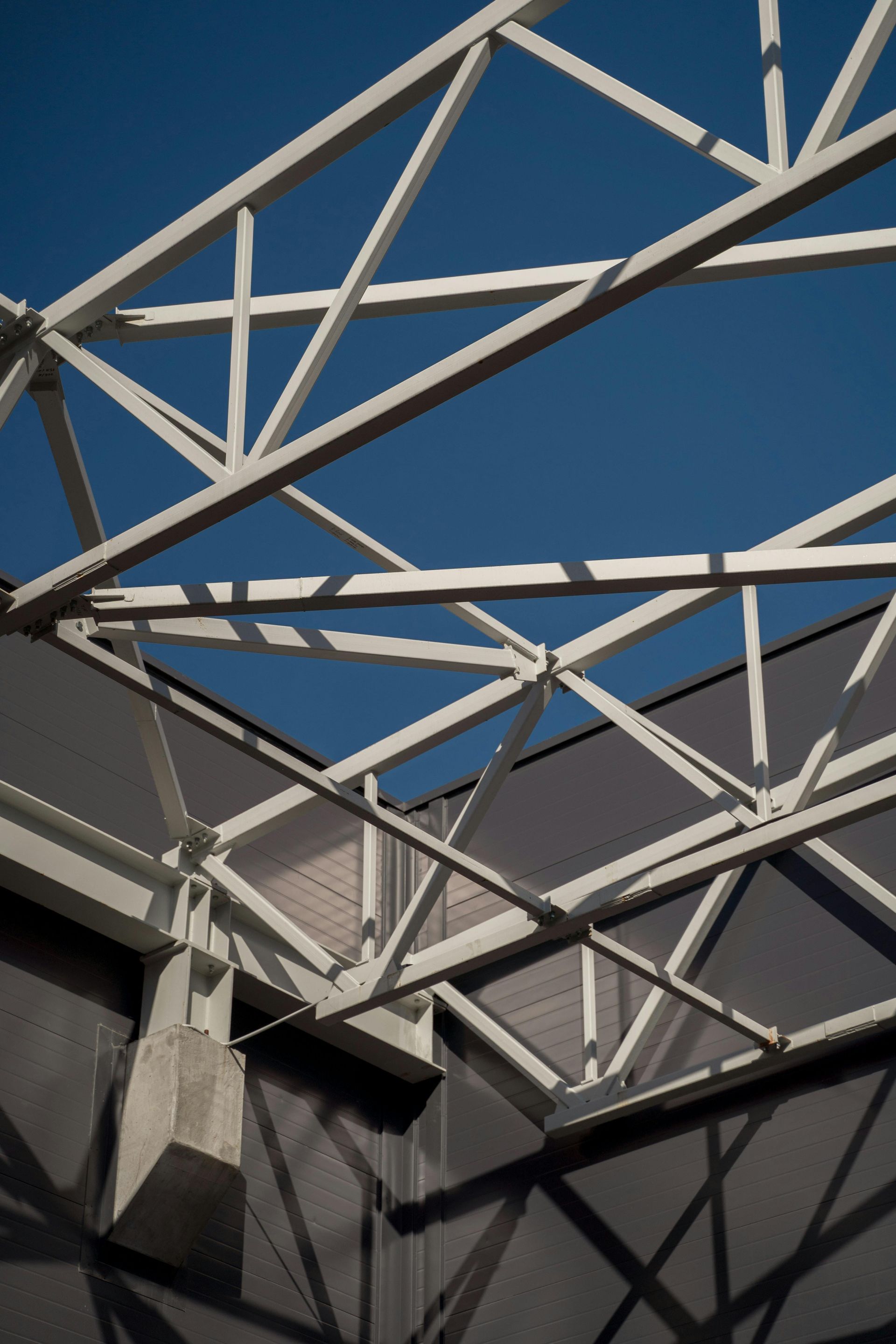 White structural steel trusses cast sharp shadows against a dark gray wall under a clear blue sky.