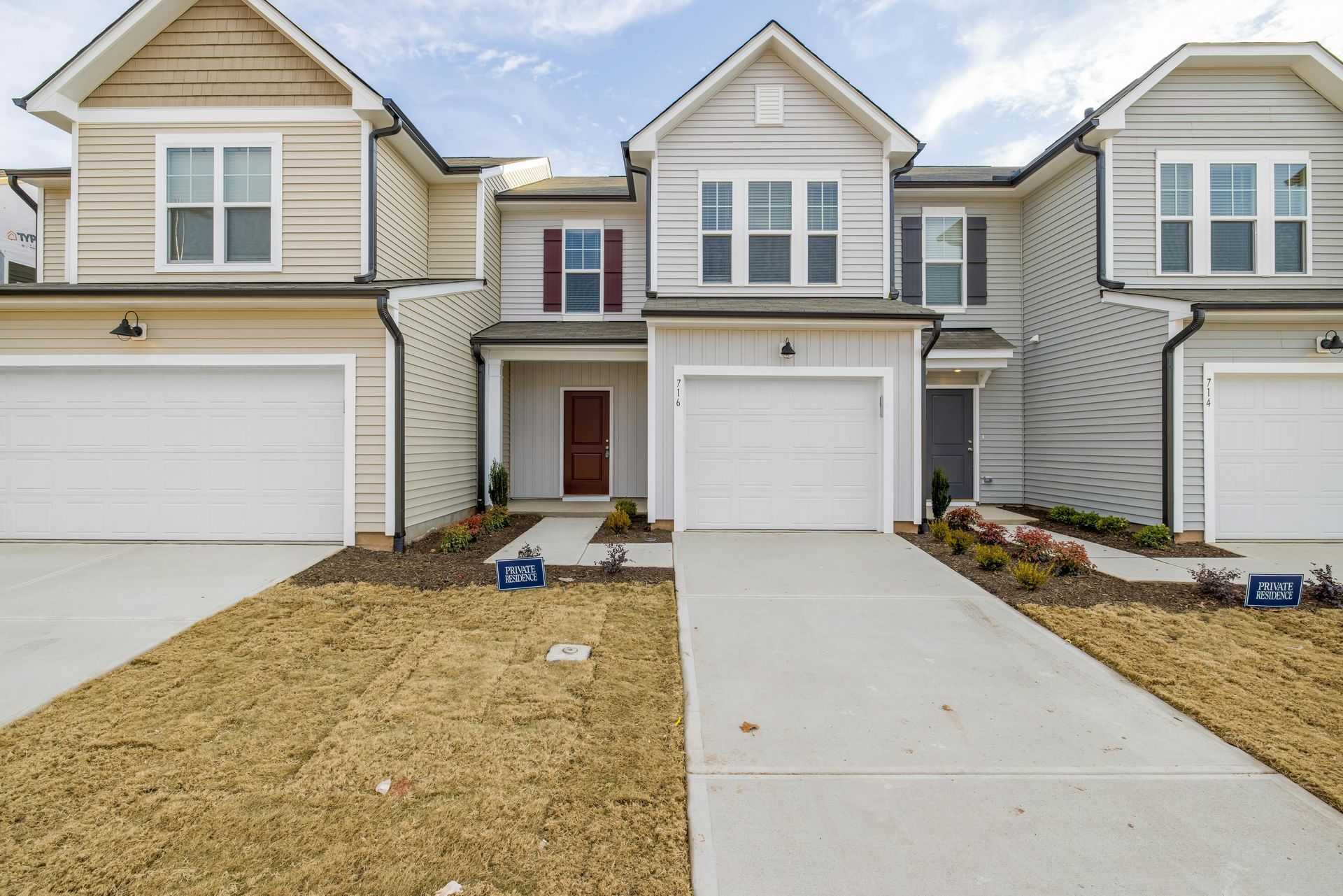 A row of three contemporary townhouses with light siding, white garage doors, and patches of dry grass in front.