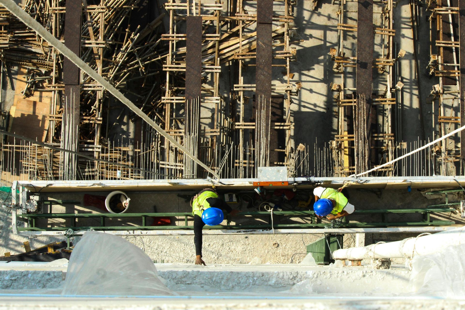 Two workers in high-visibility vests and blue hard hats work on a suspended scaffold at a construction site.