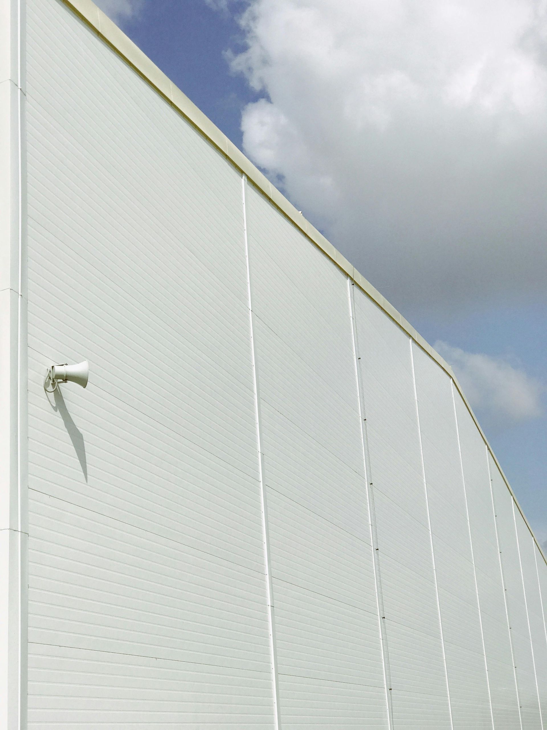 A white corrugated metal wall of a building exterior under a bright, cloudy sky with a small security camera.