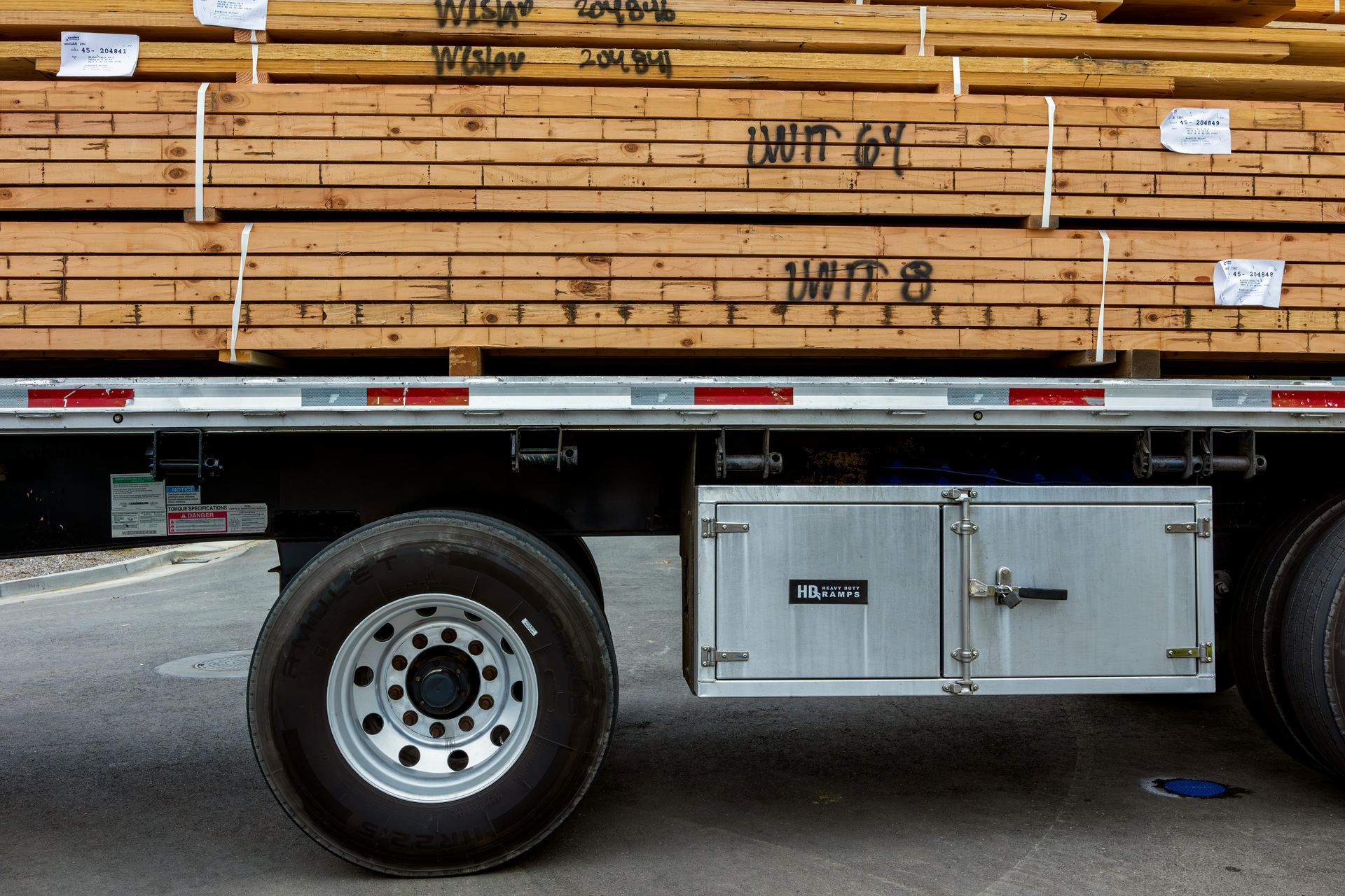 Side view of a flatbed trailer loaded with stacked lumber, featuring a metal storage box mounted on the frame.