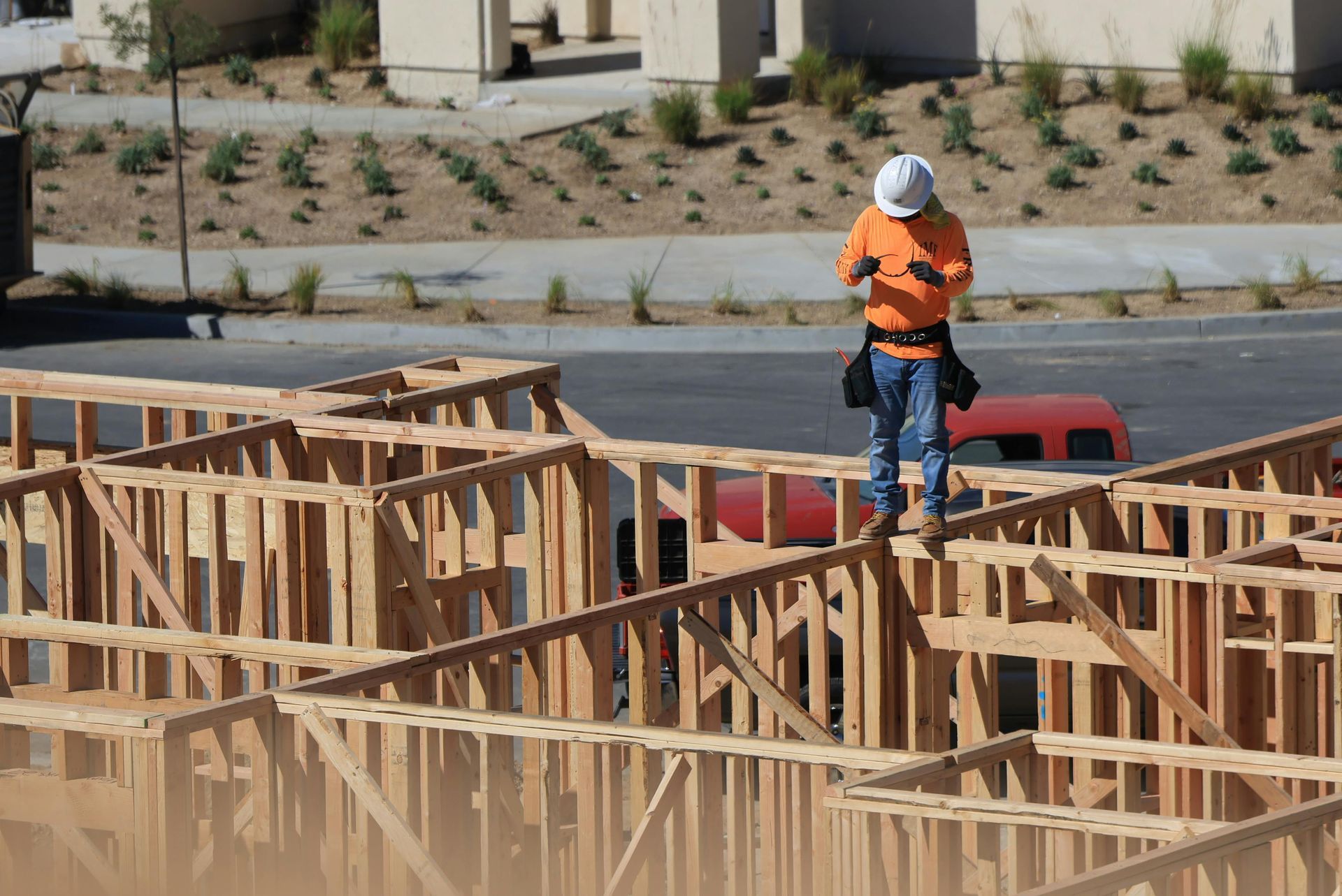 A construction worker in a hard hat and orange shirt balances on the wooden framing of a building under construction.