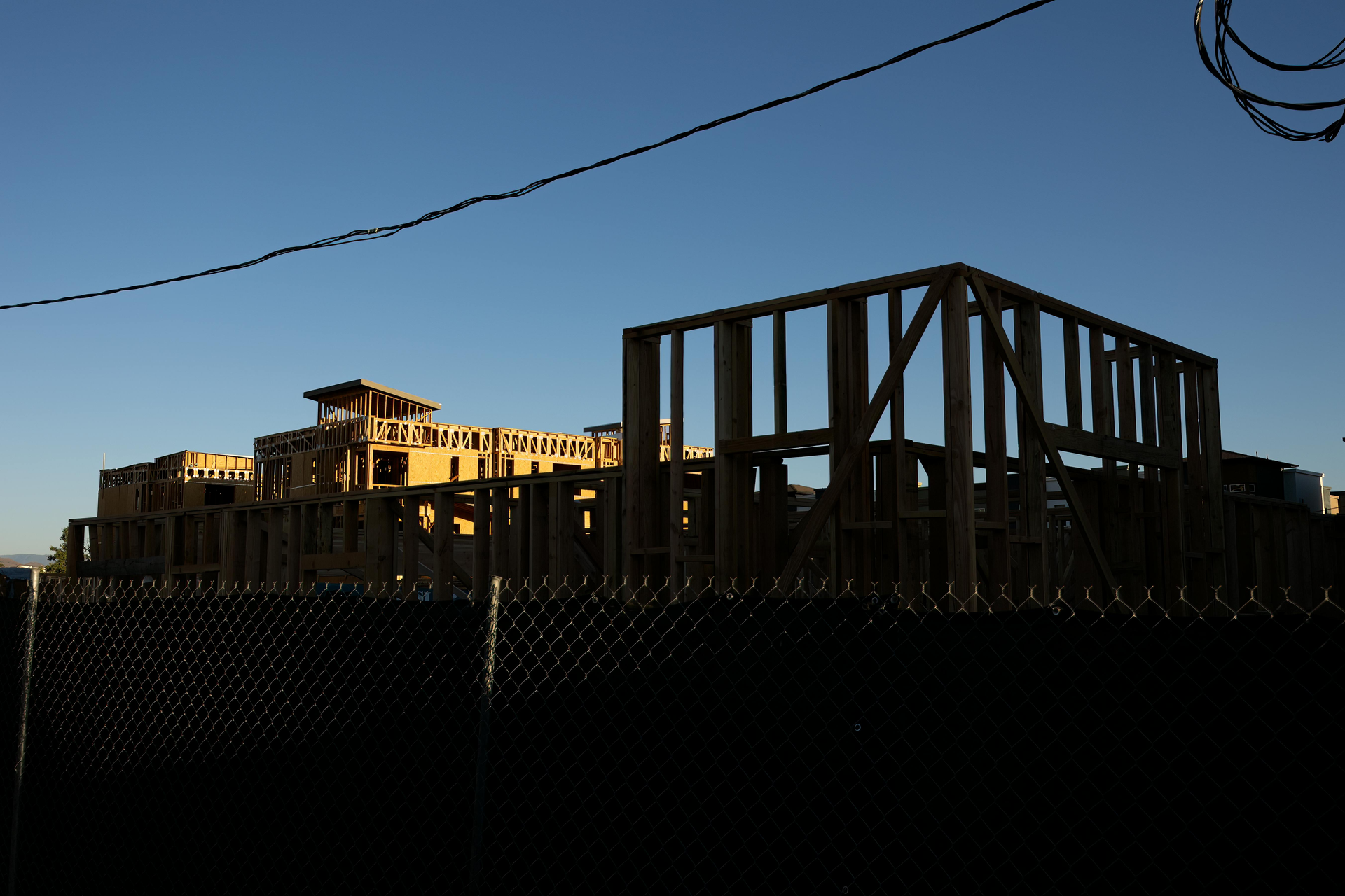 A wooden building frame under construction against a clear blue sky, viewed behind a chain-link fence.