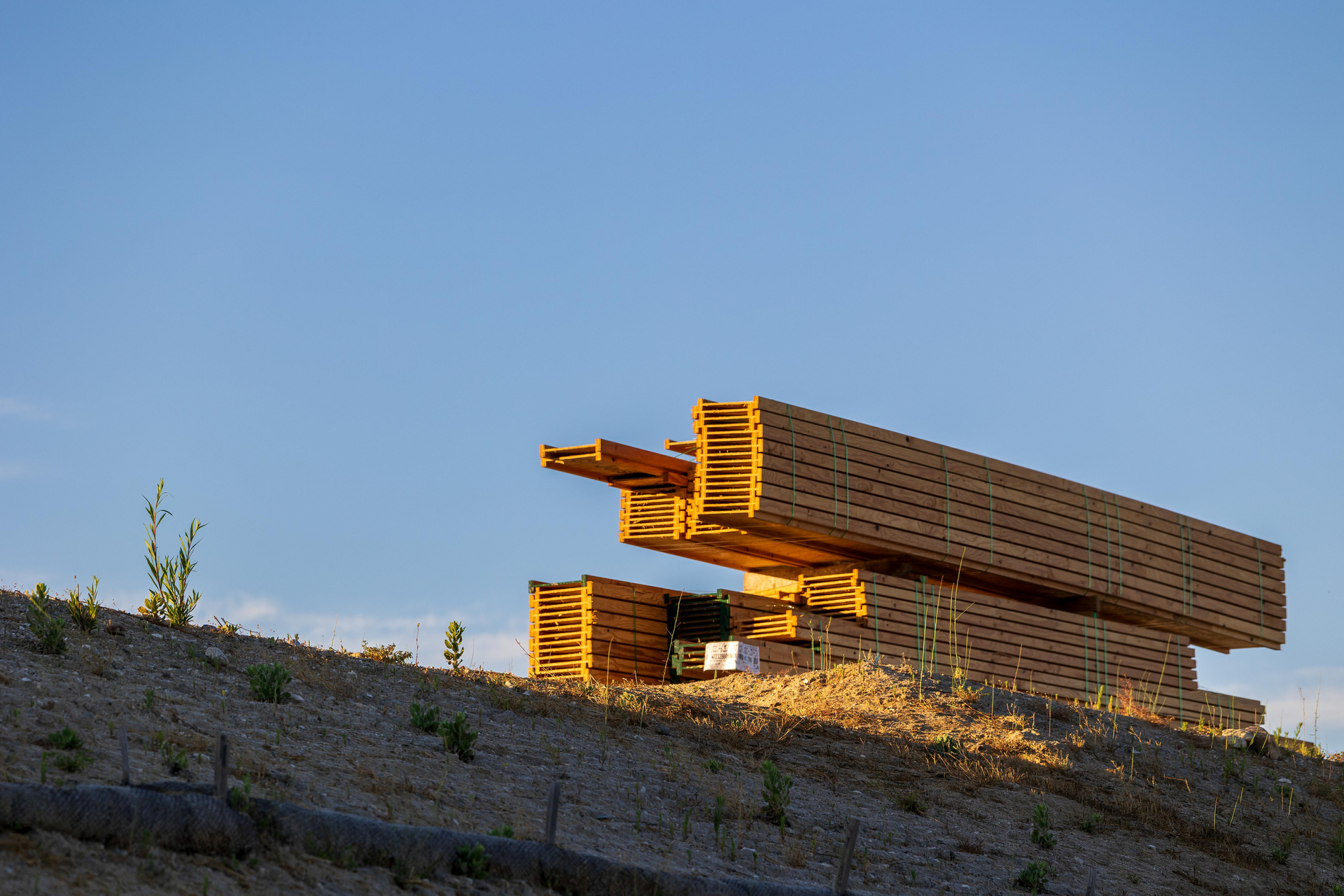 A large stack of lumber rests on a dusty, sunlit embankment against a clear blue sky.