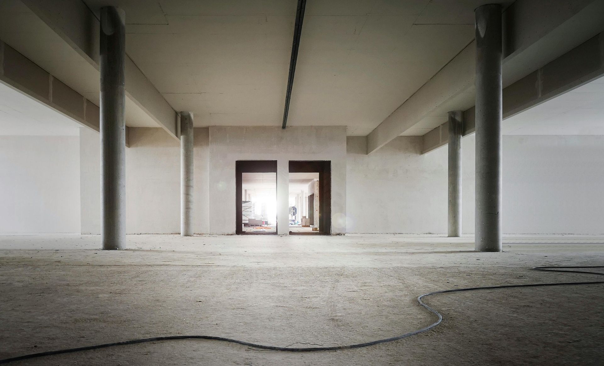 A wide, unfinished concrete room with pillars and a central doorway looking into a bright, furnished interior space.