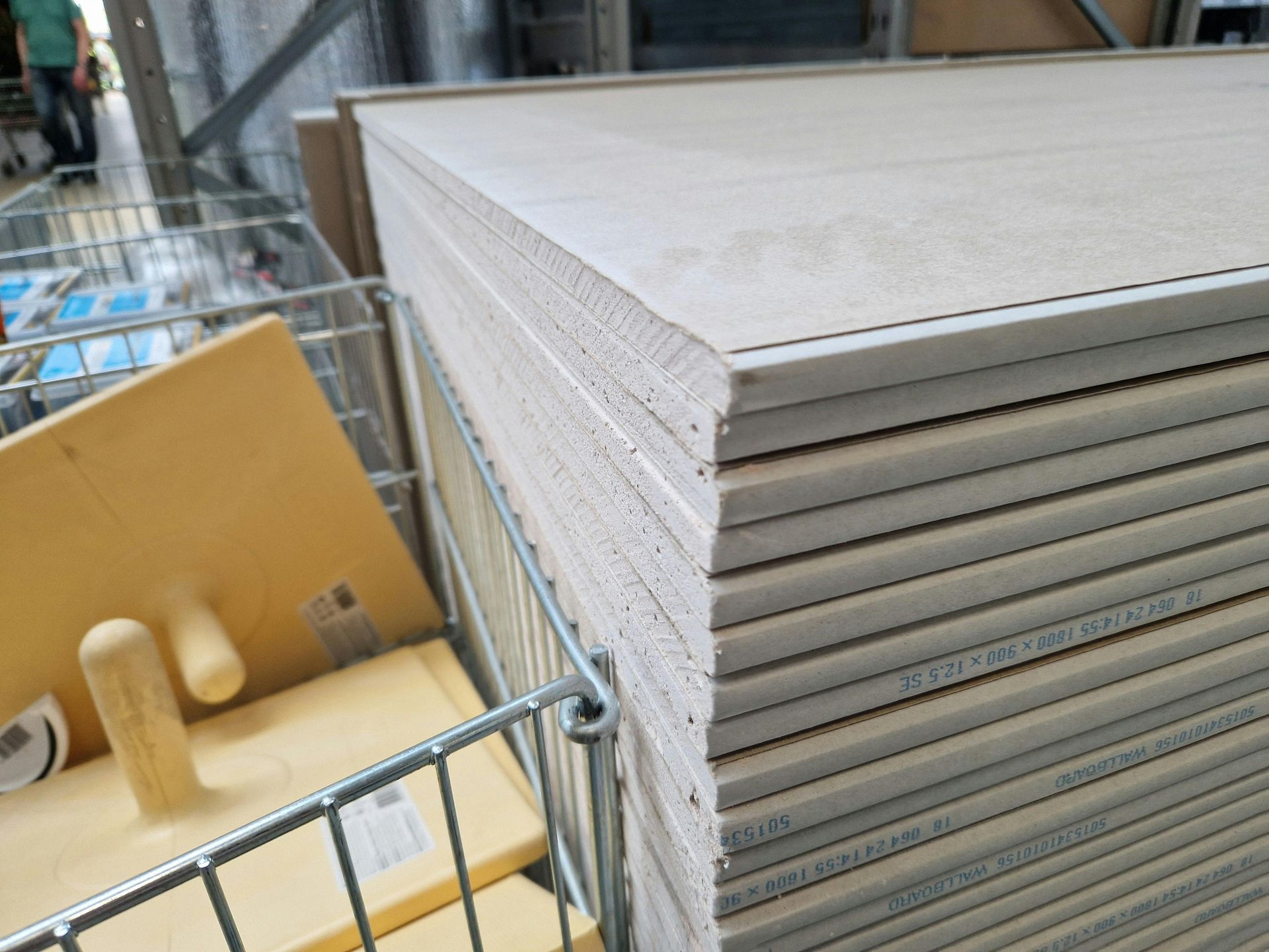 A stack of drywall panels stored in a metal retail bin next to a yellow plastic trowel.