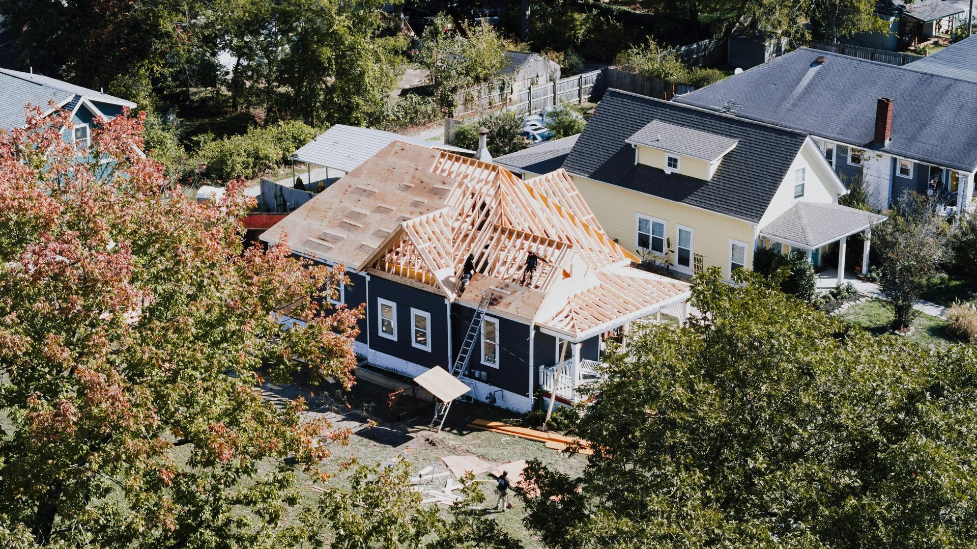 Aerial view of a blue house under construction, featuring exposed wooden roof framing, surrounded by trees and houses.