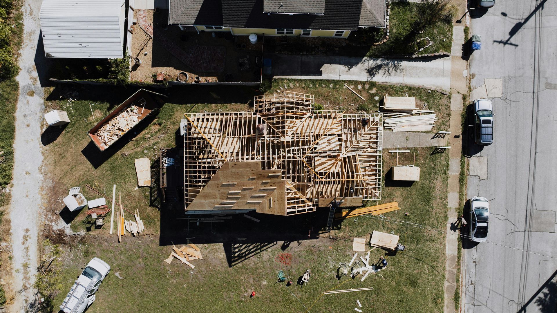 Aerial view of a house under construction with exposed wooden framing and a partial roof, set on a grass lot with vehicles.