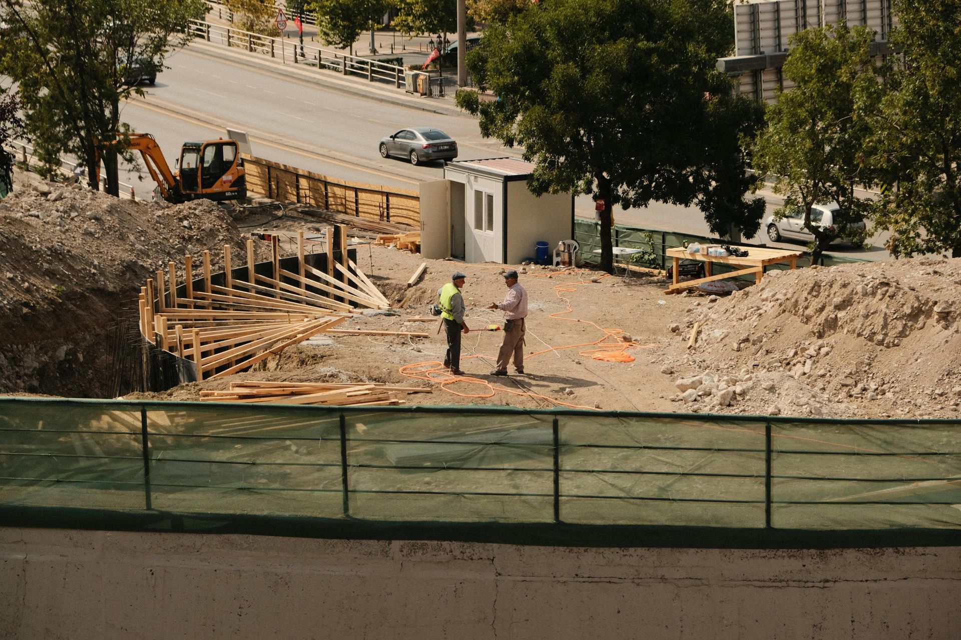 Two people in work clothing stand talking on a construction site with piles of dirt.