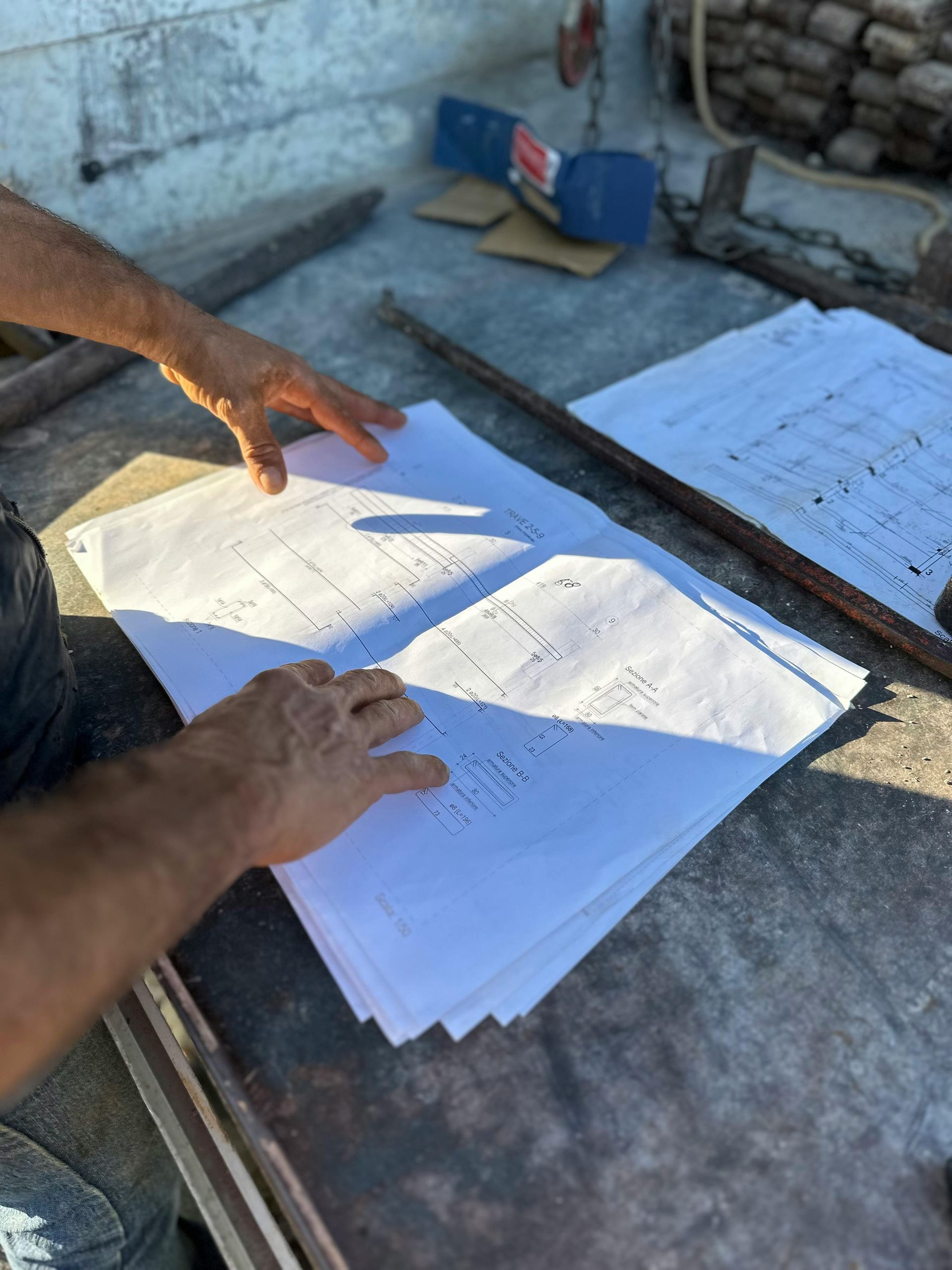 A person's hands review printed construction blueprints spread out on a flat, concrete-like surface at a work site.