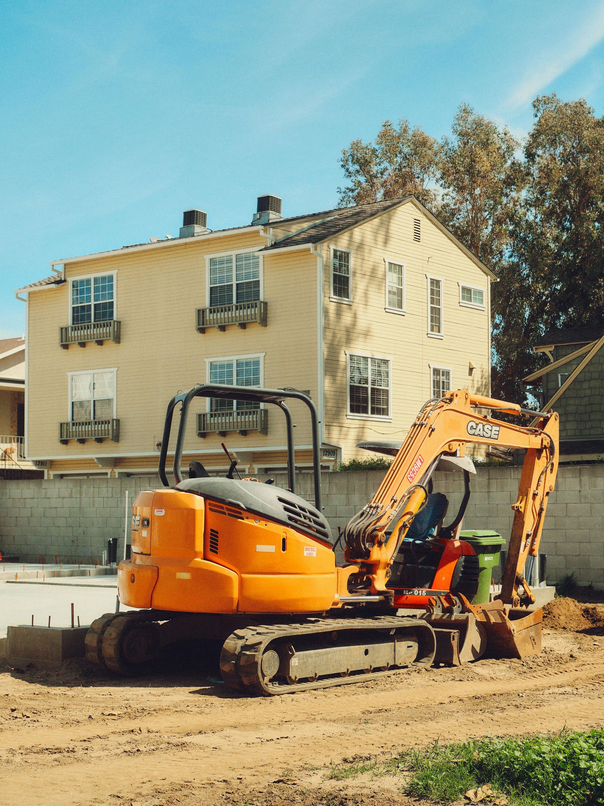 An orange excavator sits on a dirt lot in front of a yellow two-story house with window boxes.