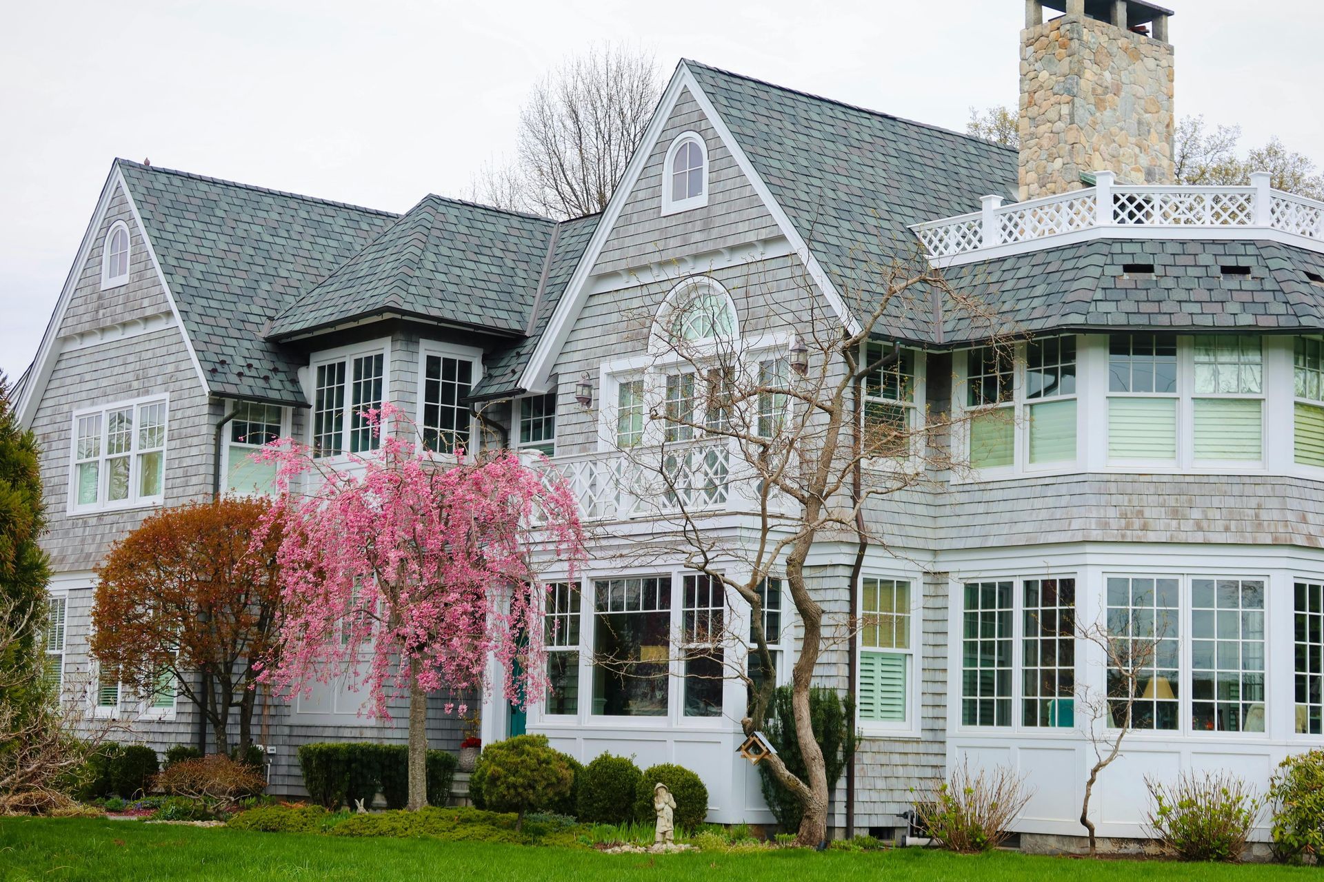 A large, two-story house with gray shingles, a steep roof, a stone chimney, and a small pink flowering tree.