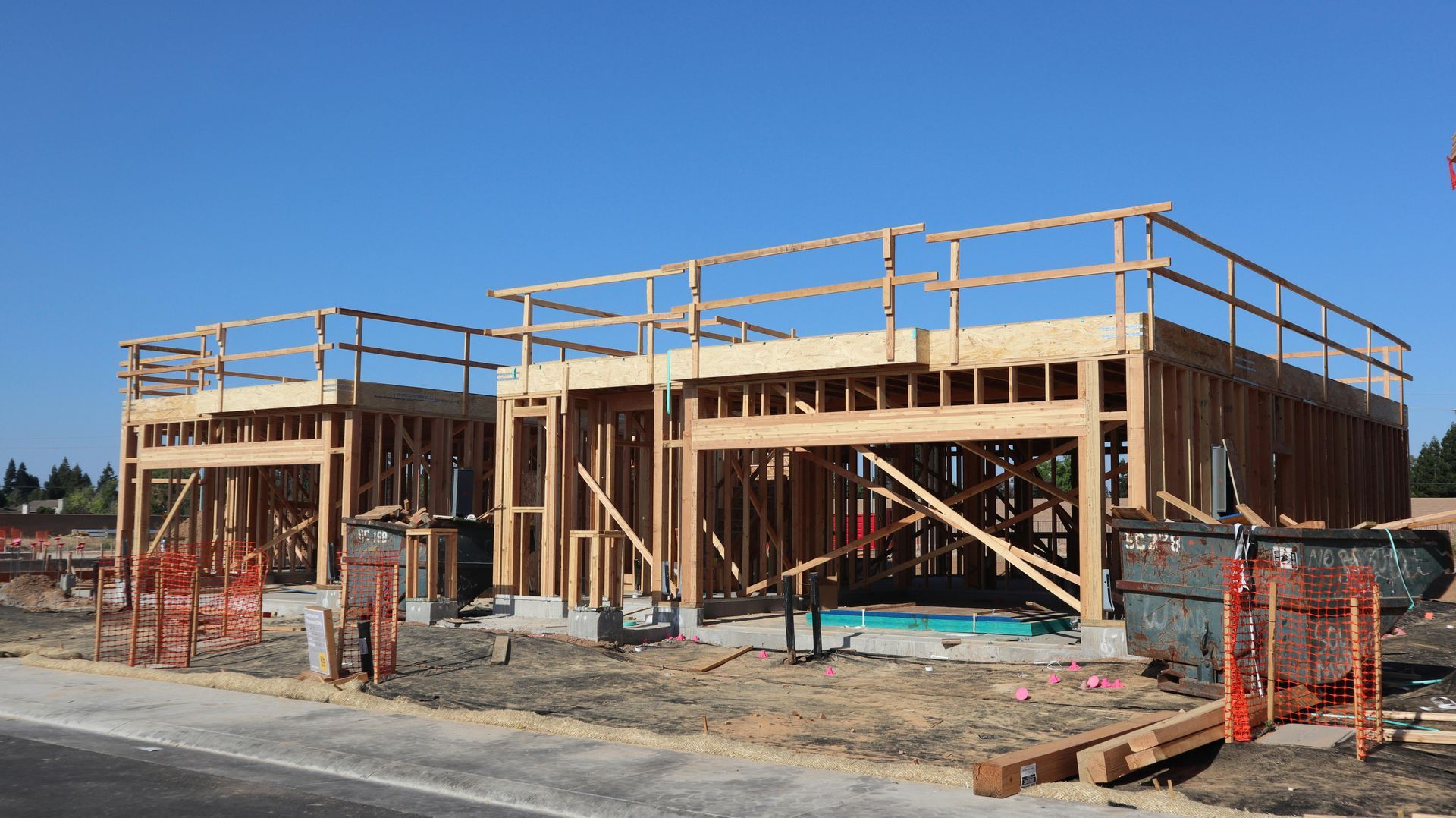 Two wooden building frames under construction with temporary safety railings and a dumpster on a dirt lot.