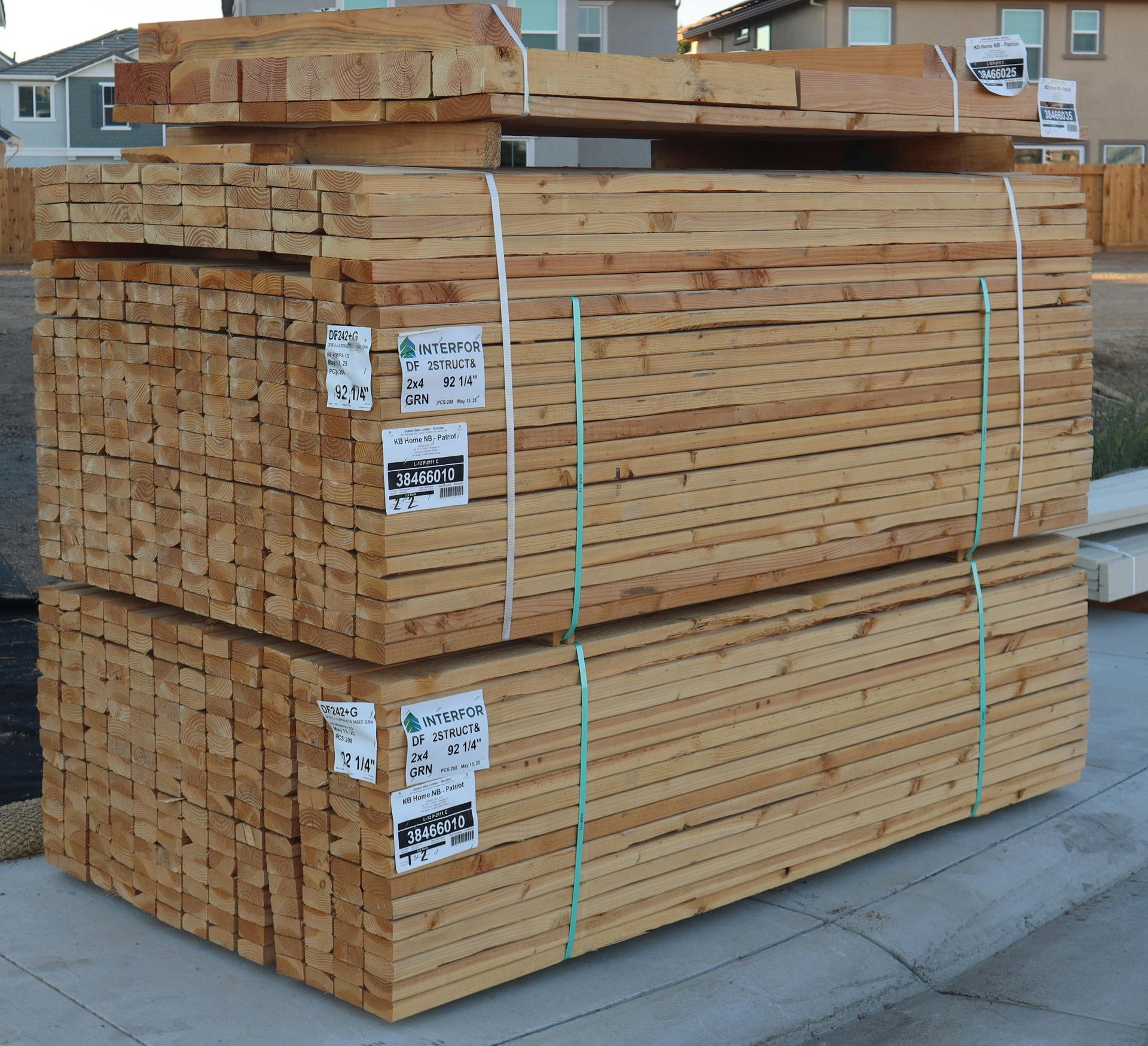A large stack of lumber wrapped in plastic bands sits on a concrete surface in a residential neighborhood.
