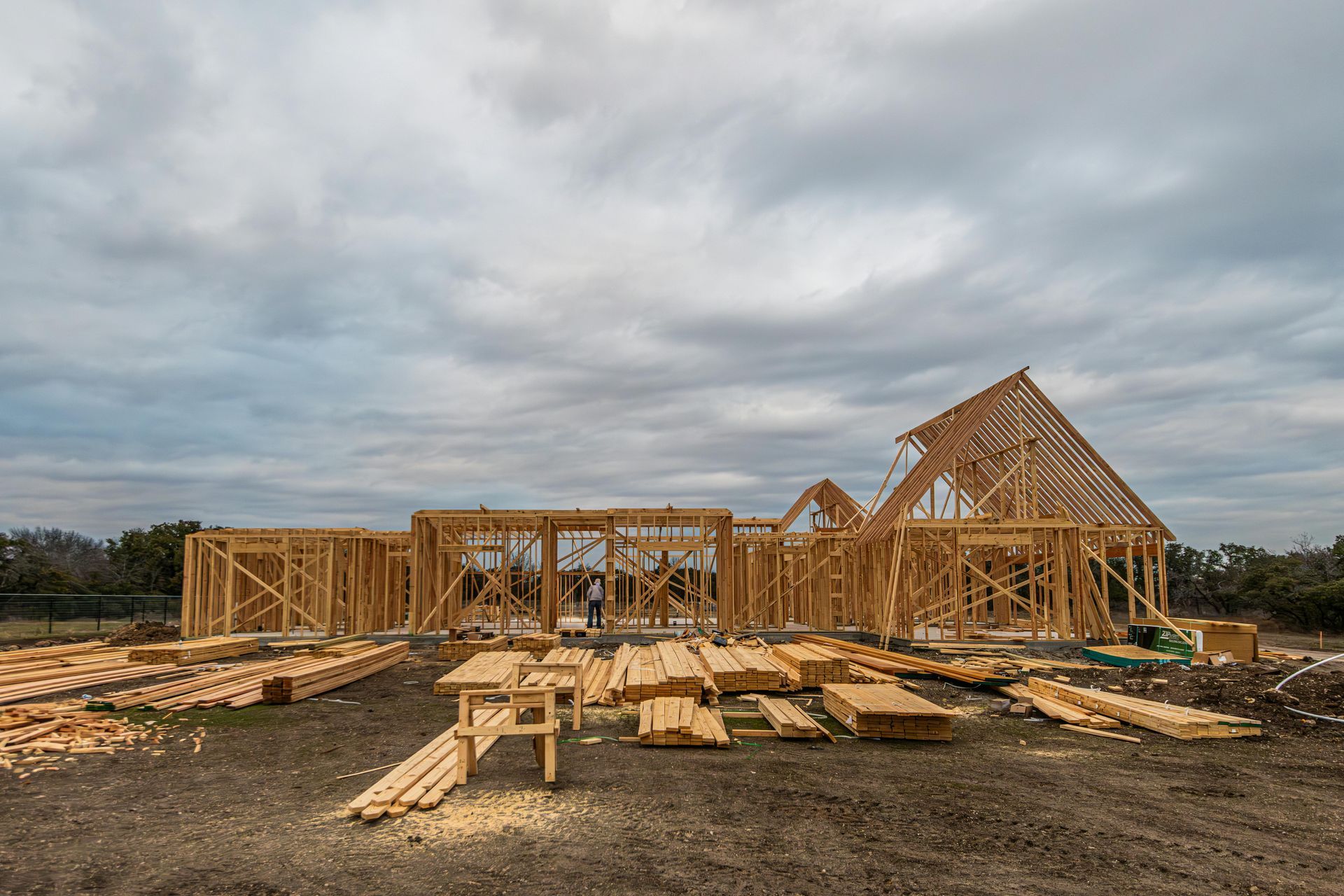 Wooden frame of a house under construction on a dirt lot beneath a cloudy sky, with stacks of lumber in the foreground.