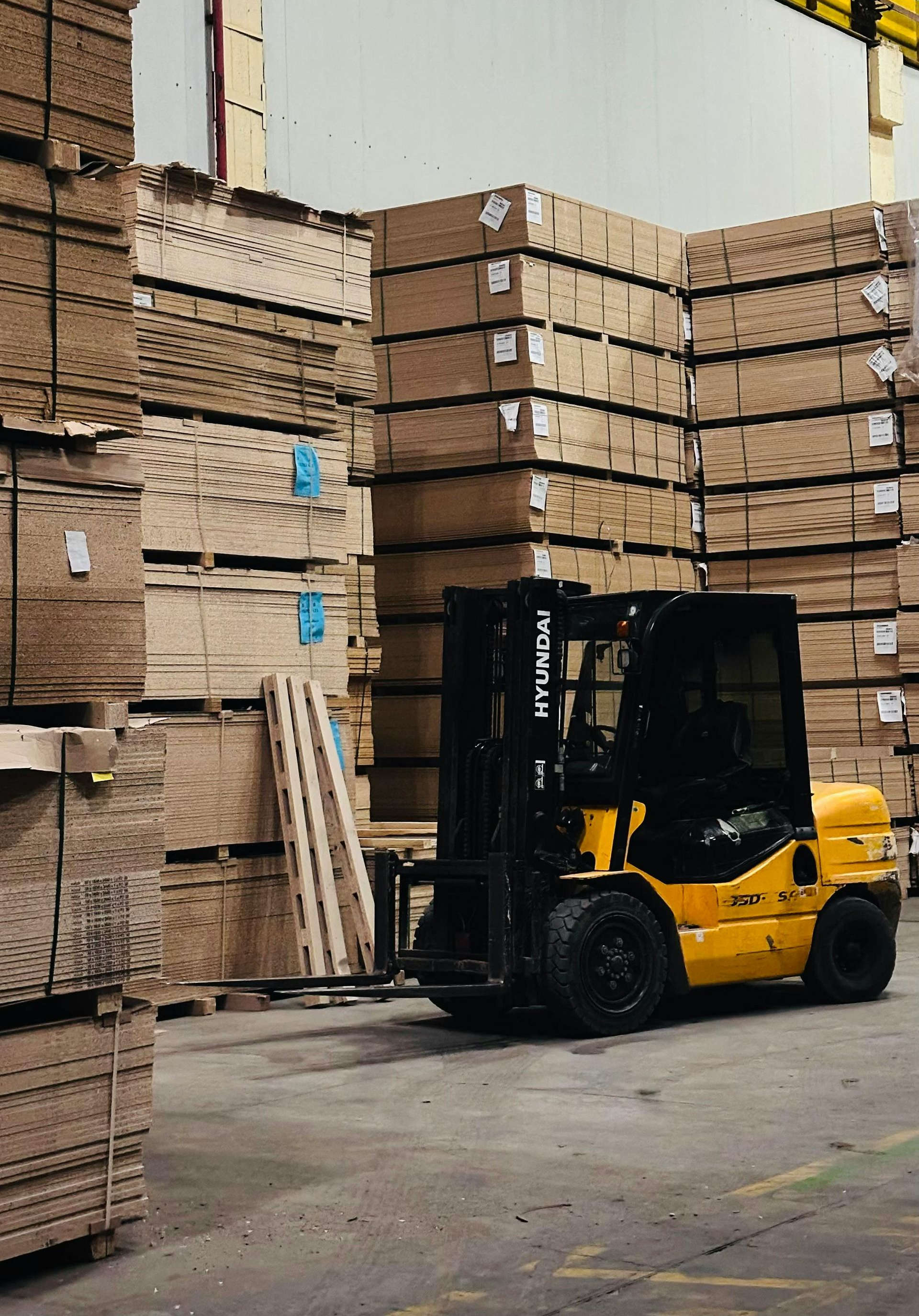 A yellow forklift operates in a warehouse aisle lined with tall, stacked pallets of wood panels.