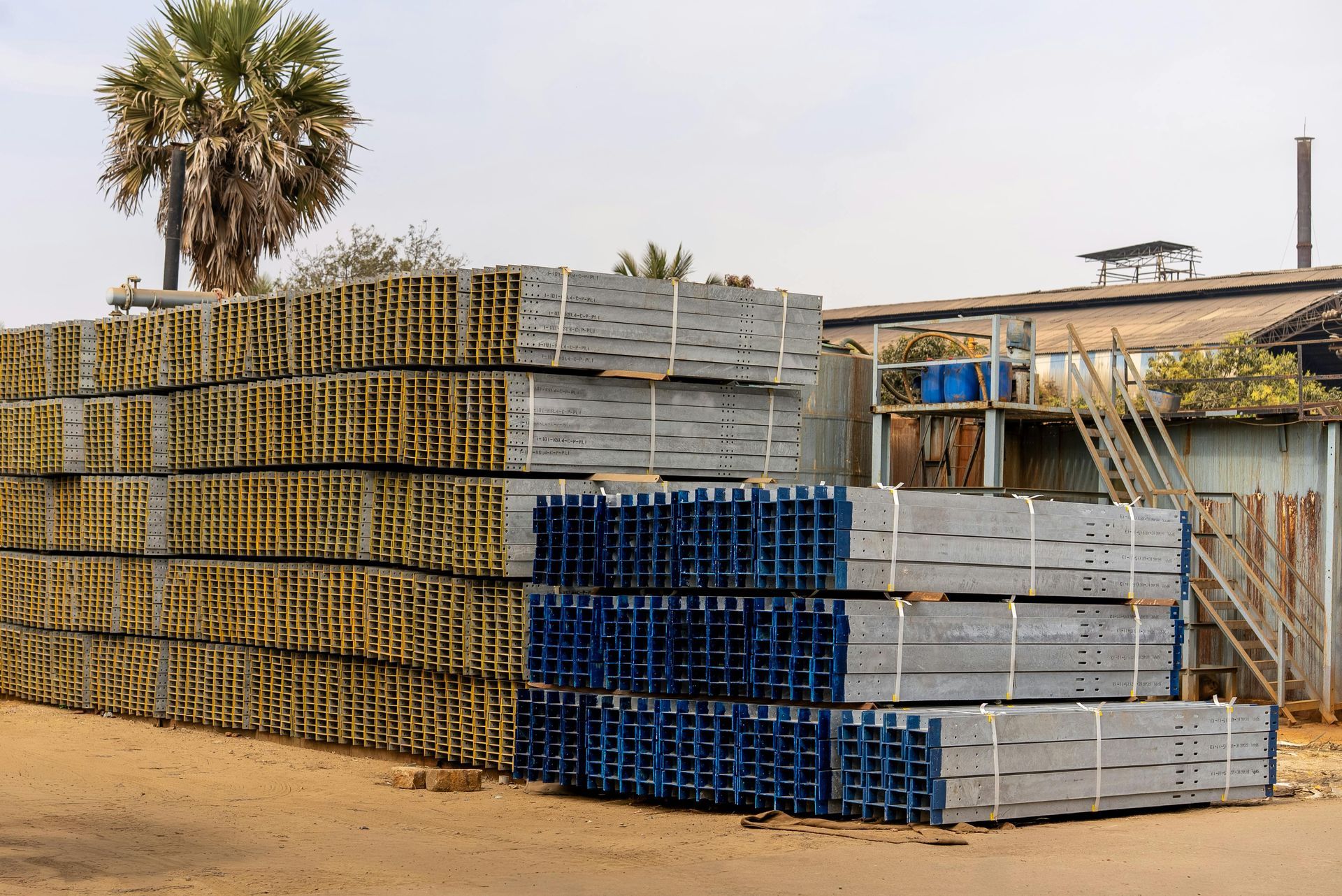 Large stacks of industrial metal beams and girders stored outdoors near a building under a bright sky.