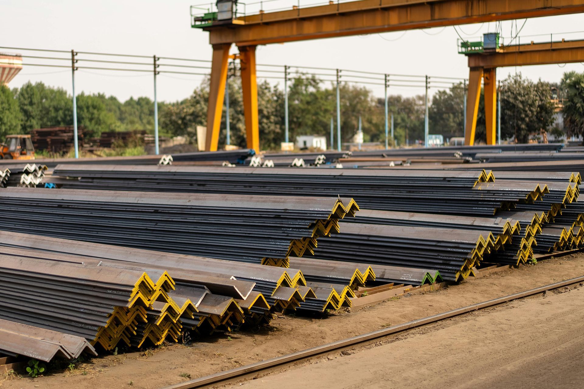 A large outdoor industrial yard stacked with rows of dark gray steel angle bars under a yellow overhead crane gantry.