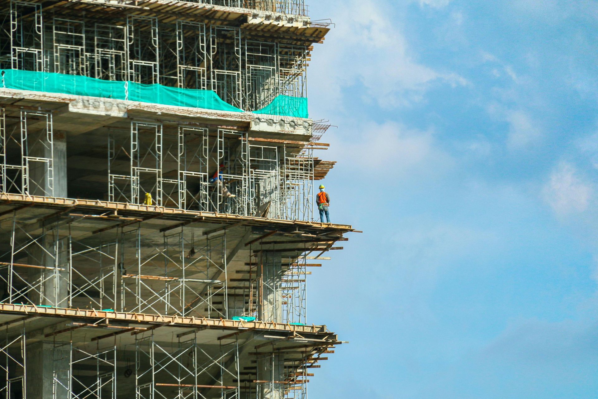 A construction worker stands on the edge of an unfinished, scaffold-covered high-rise building against a blue sky.