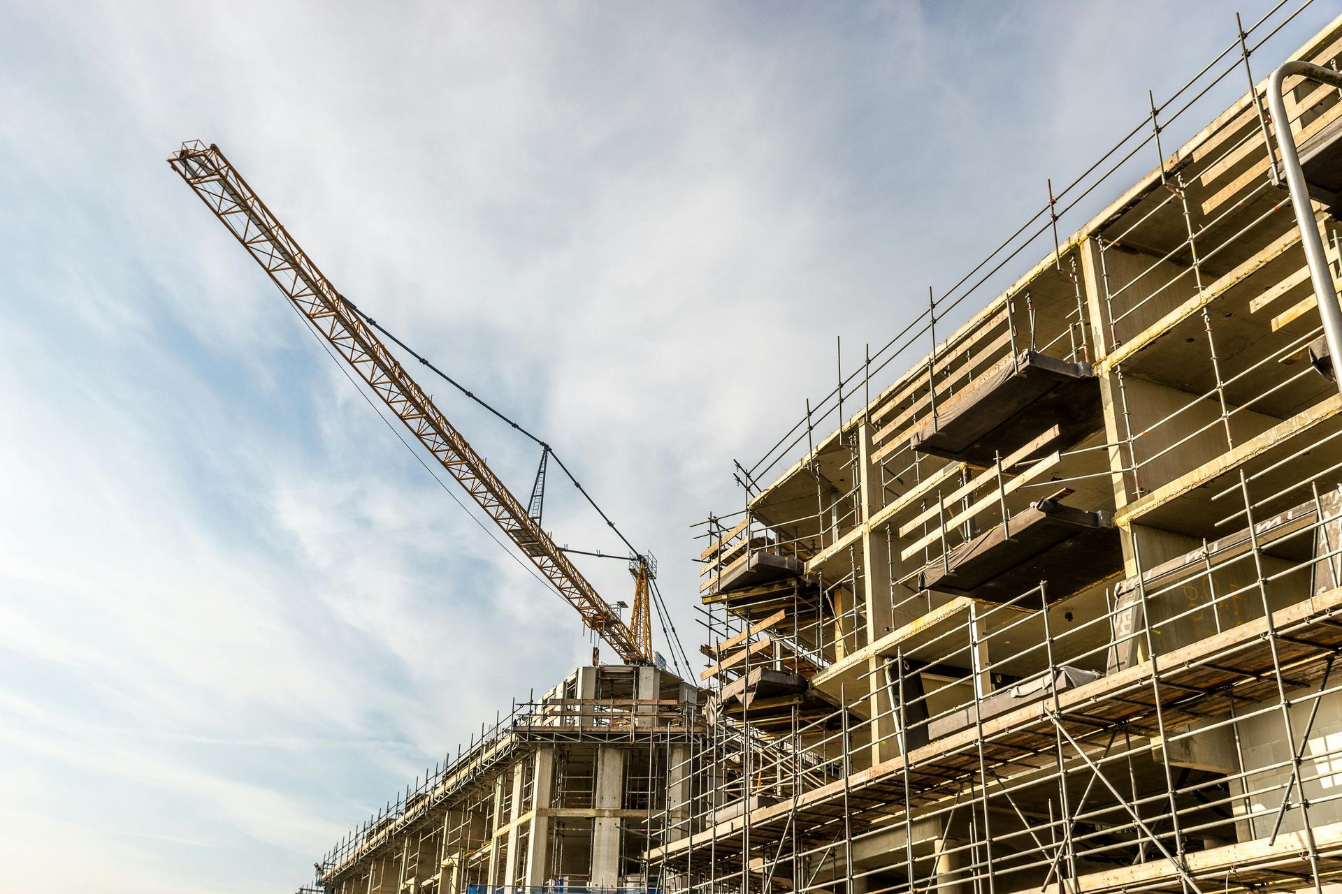 A multi-story building under construction with metal scaffolding and a large crane against a bright, cloudy sky.