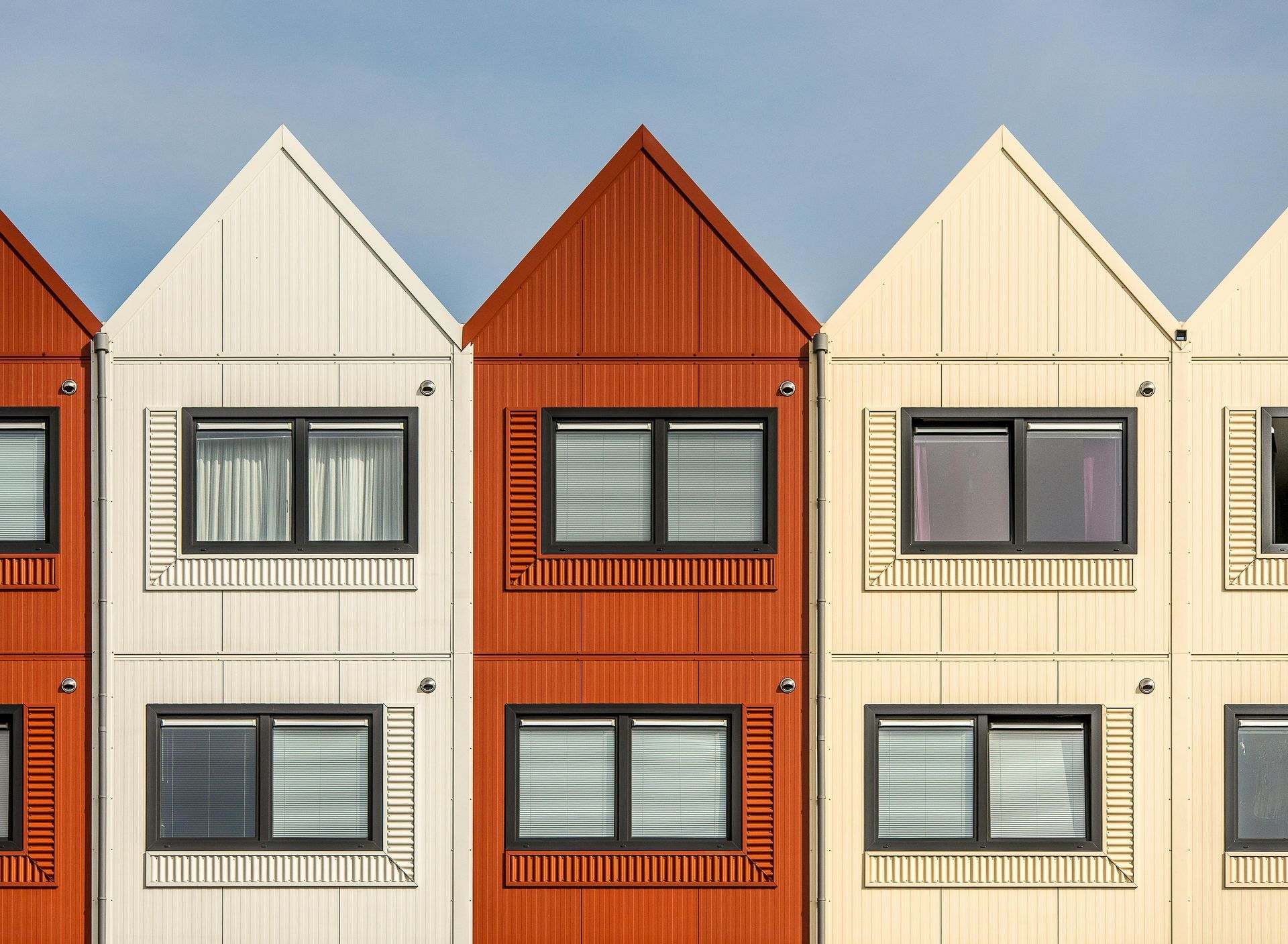 A row of modern, narrow houses with triangular roofs in alternating colors of red, white, and tan against a blue sky.