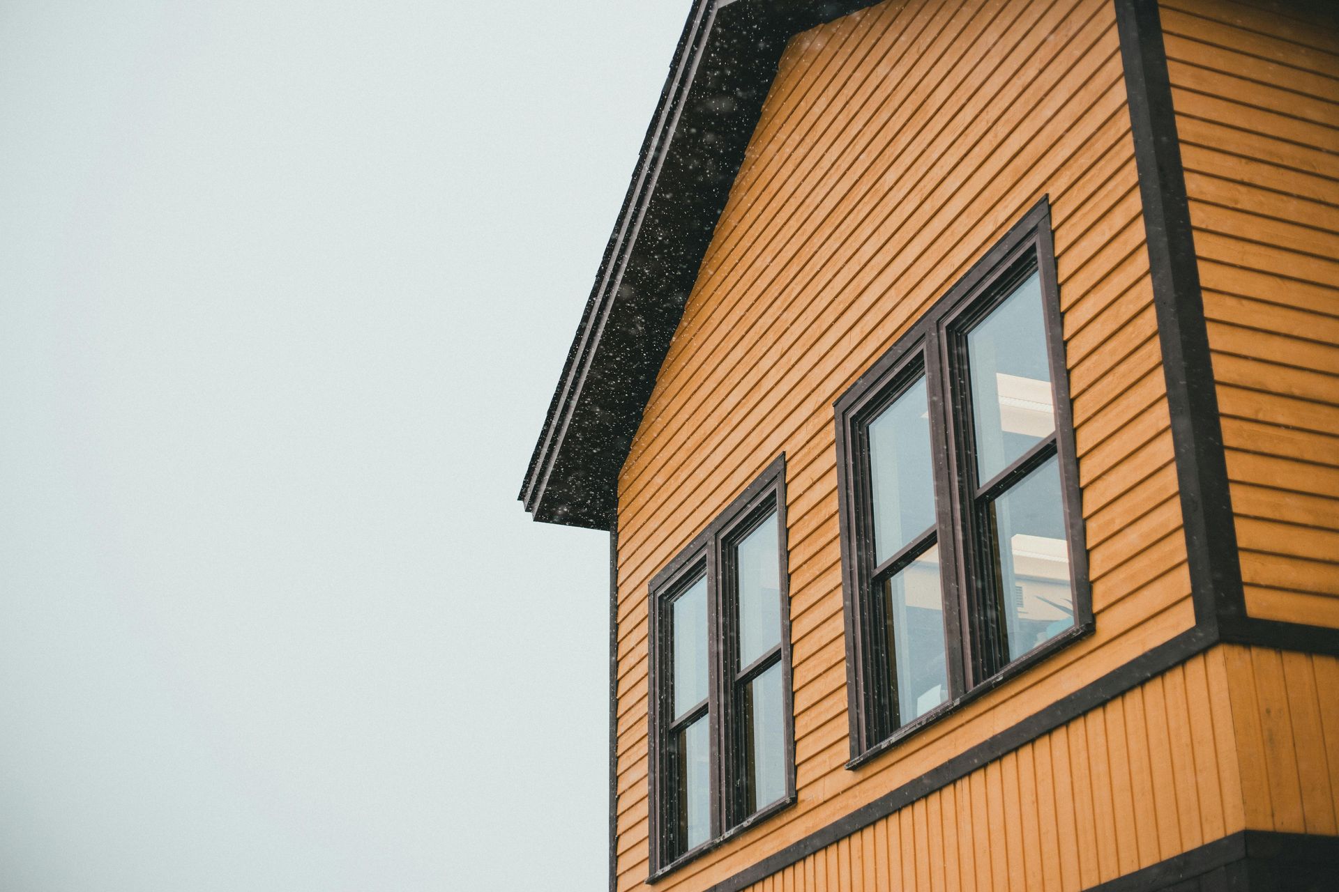 Low angle shot of a mustard-yellow house with dark trim and two windows against a pale, overcast sky.