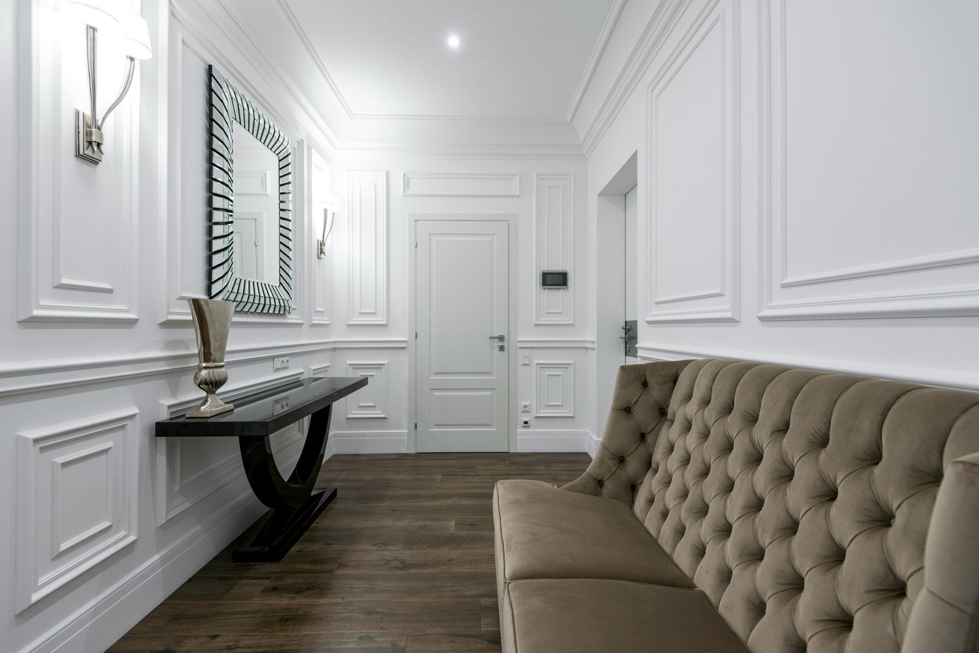 A hallway with white wainscoting, a tufted brown bench, a black console table, and a mirror on a dark wooden floor.