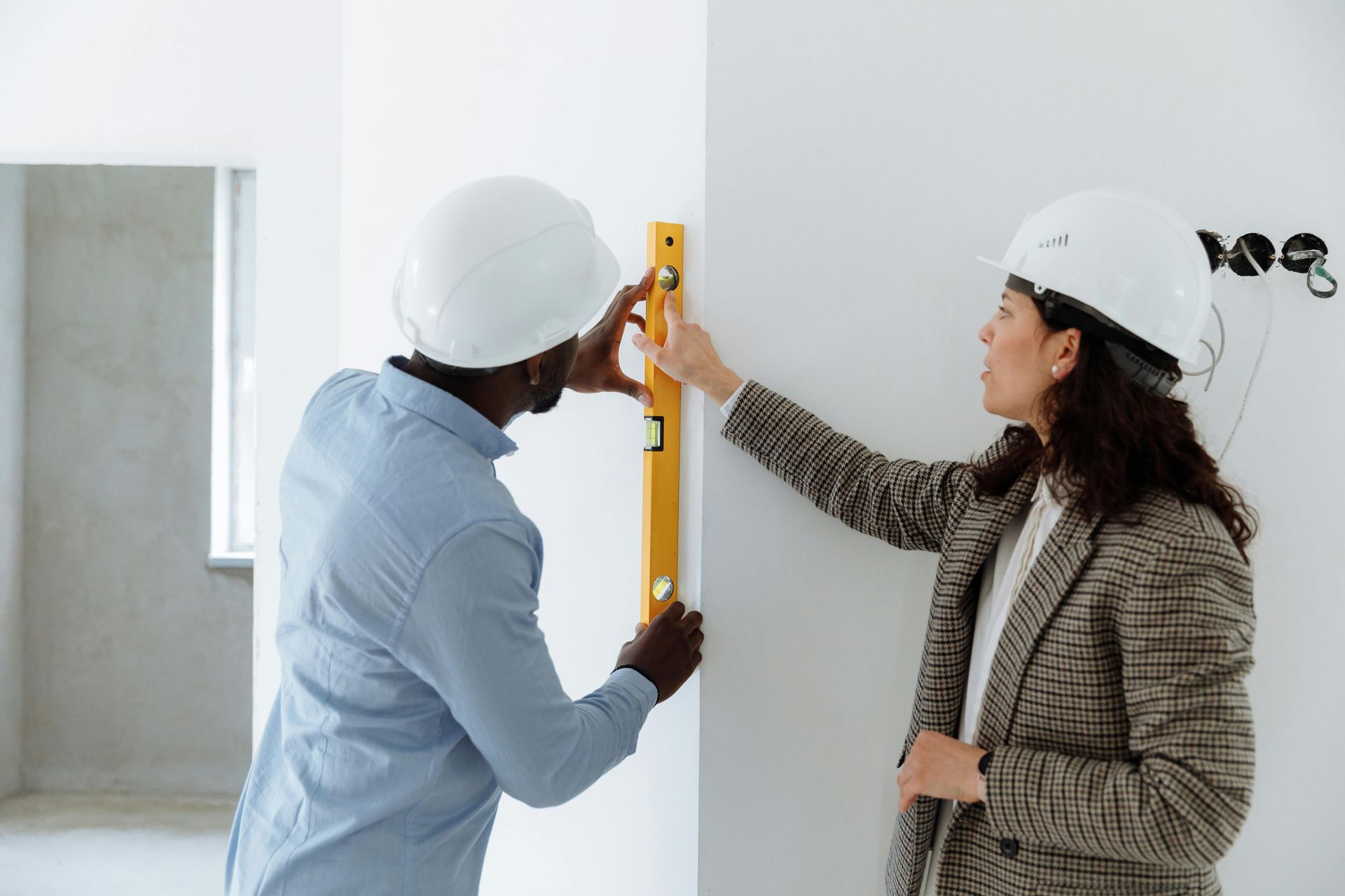 Two people wearing hard hats use a yellow spirit level to check the vertical alignment of a wall in a building project.