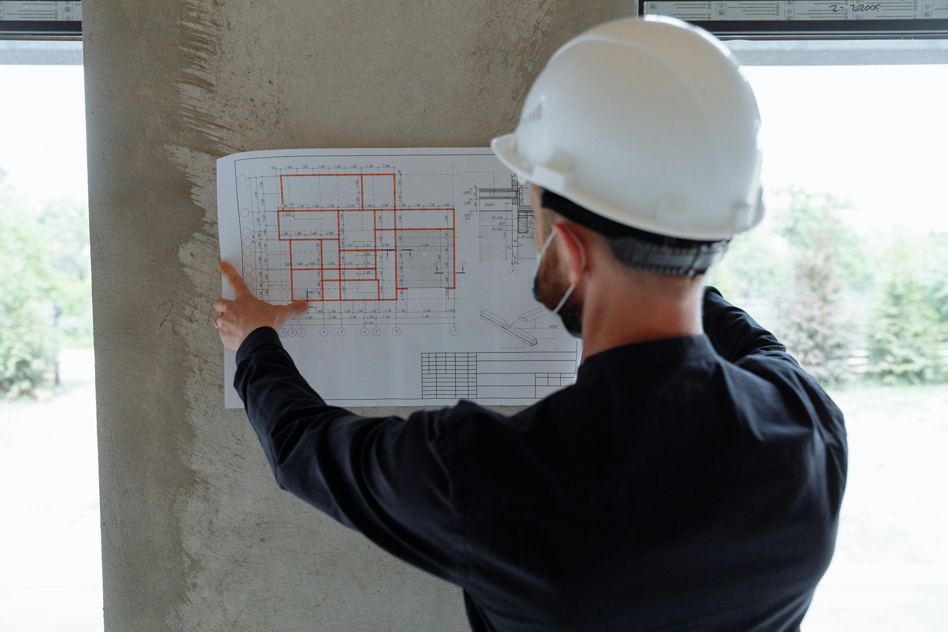 A person wearing a white hard hat and a black shirt examines a floor plan blueprint attached to a concrete pillar.