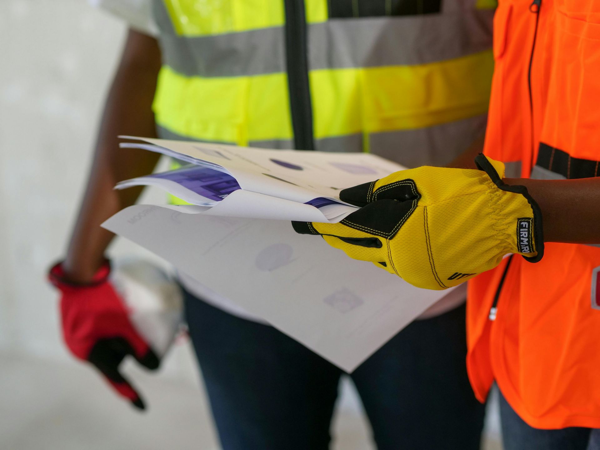 Two construction workers wearing high-visibility vests and safety gloves examine a set of building plans.