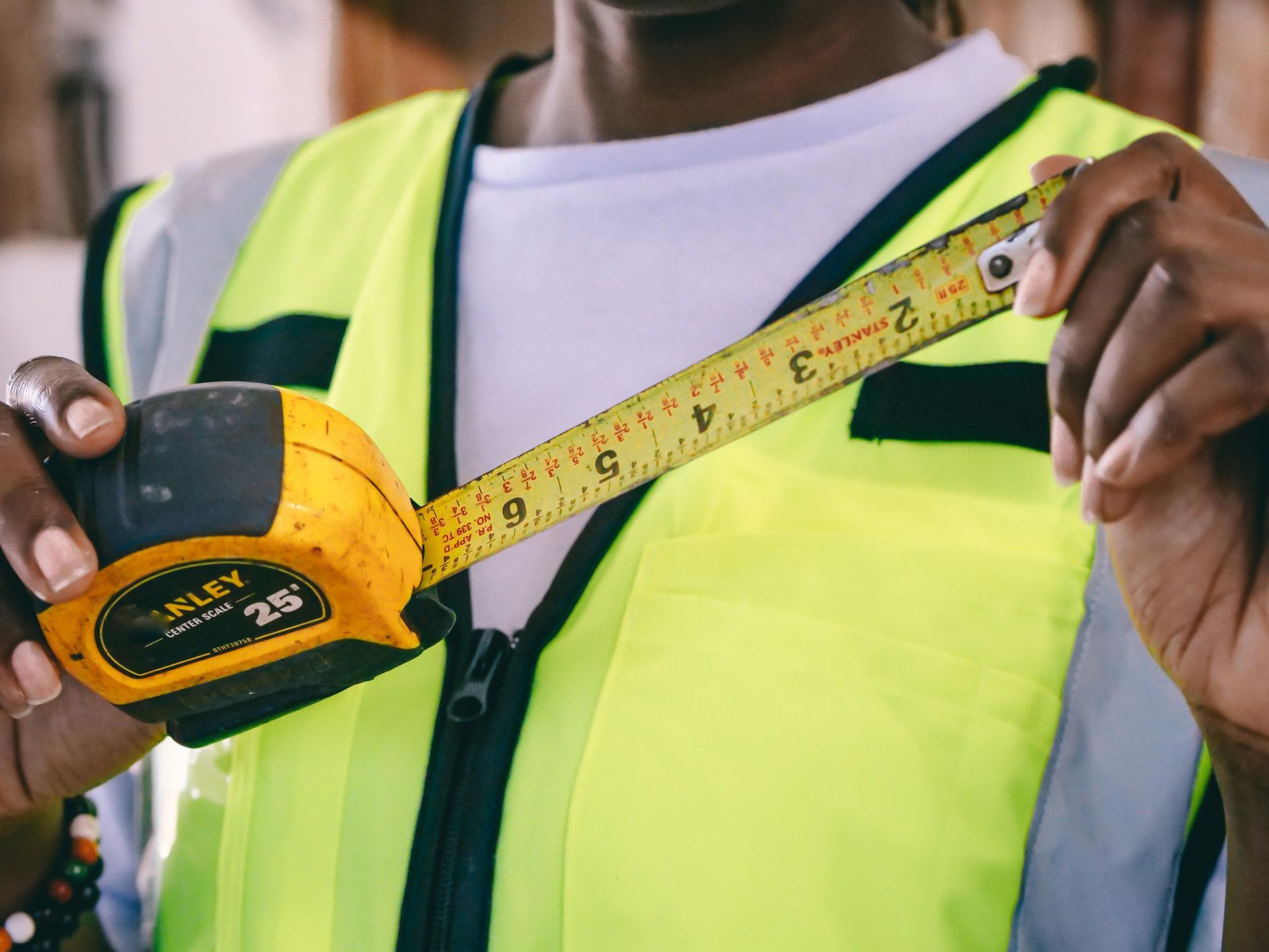 A person in a high-visibility yellow vest holds and reads a yellow Stanley 25-foot tape measure.