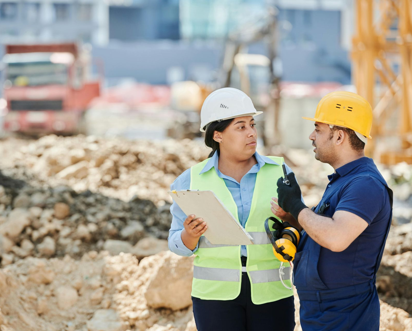 Two construction professionals in hard hats and vests discuss plans on a worksite.