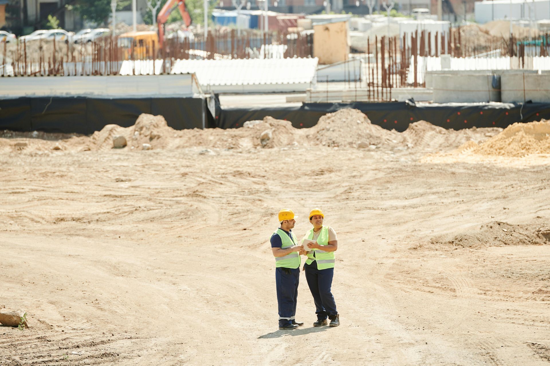 Two workers in high-visibility vests and yellow hard hats stand on a large dirt construction site, talking.
