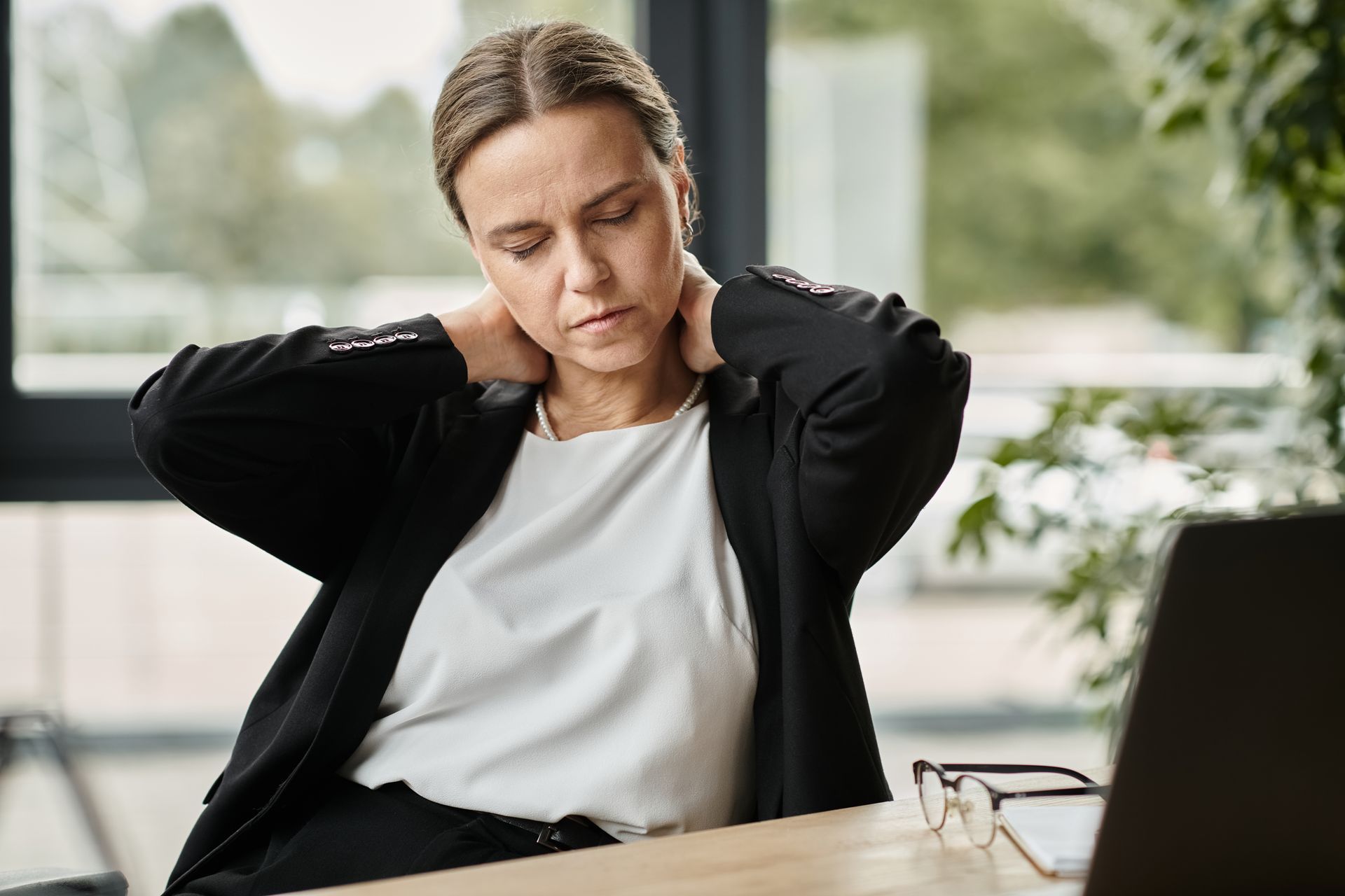 A woman is sitting at a desk with a laptop and holding her neck.