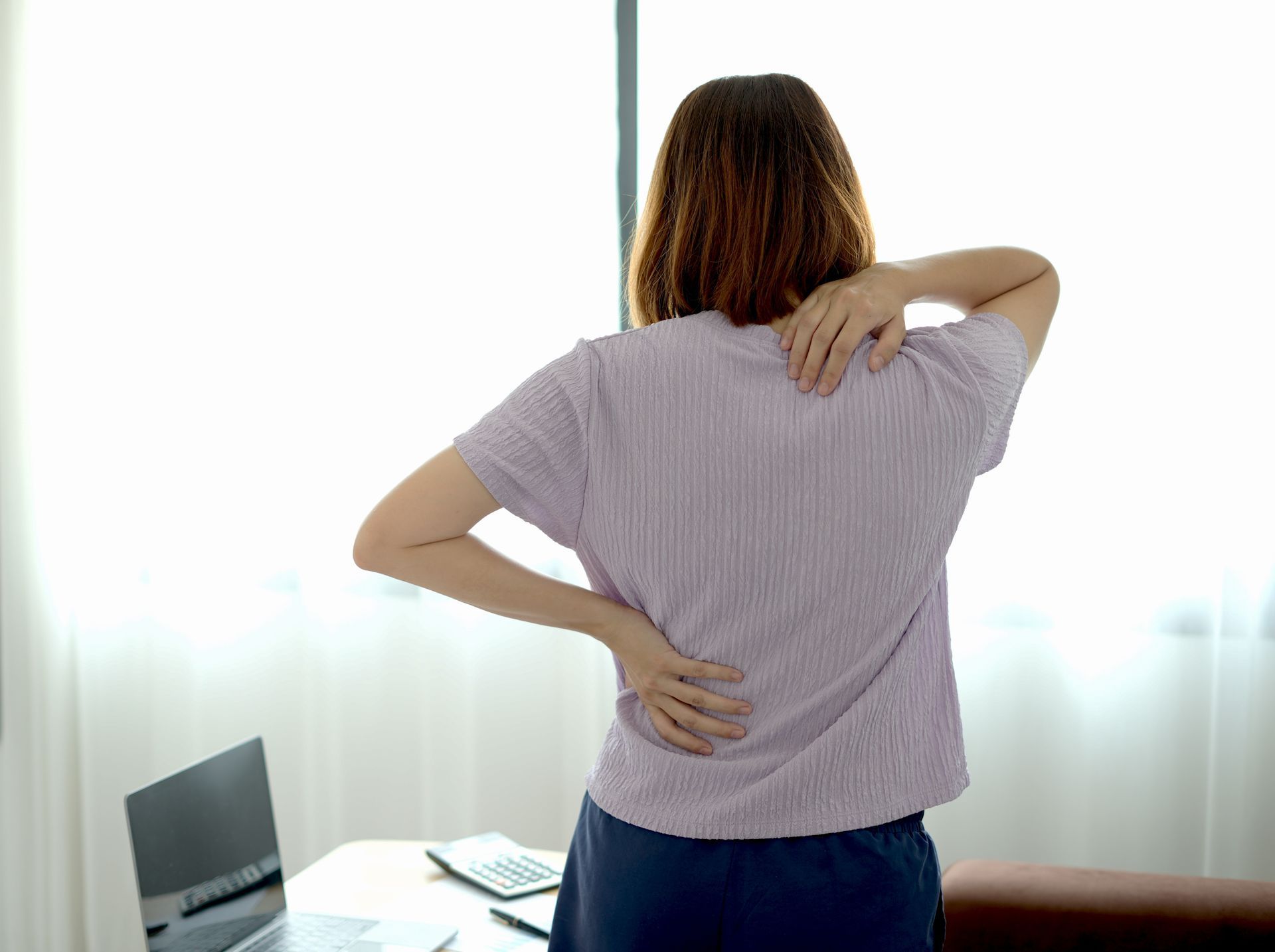 A woman is standing in front of a window holding her back in pain.