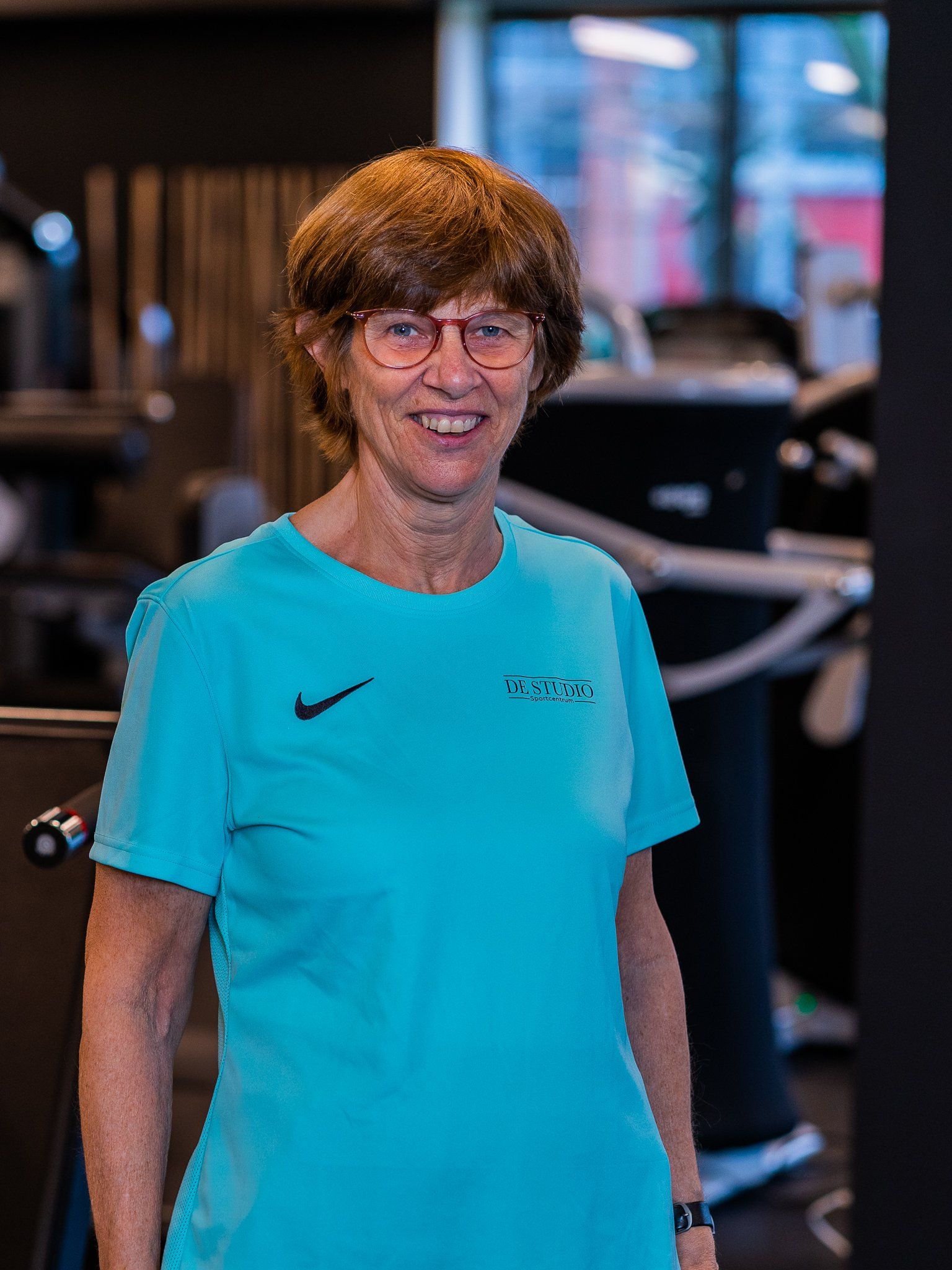 Een vrouw met een bril en een blauw shirt staat in een sportschool.