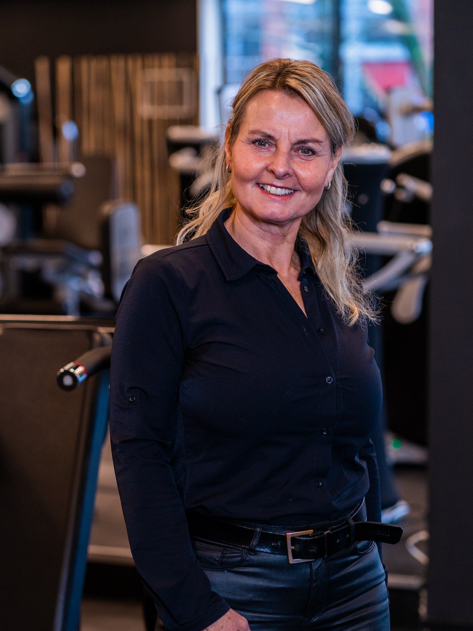 Een vrouw staat in een sportschool, draagt ​​een zwart shirt en glimlacht.